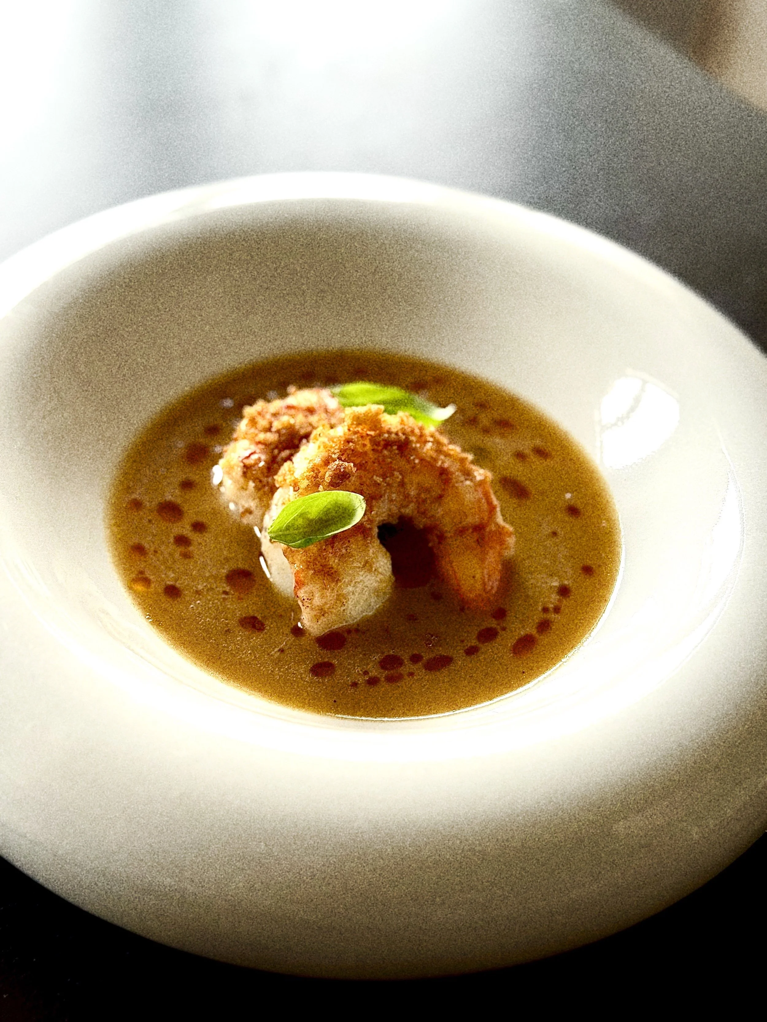 A white bowl of savory sauce topped with shrimp, fresh green herbs, and a drizzle of oil.