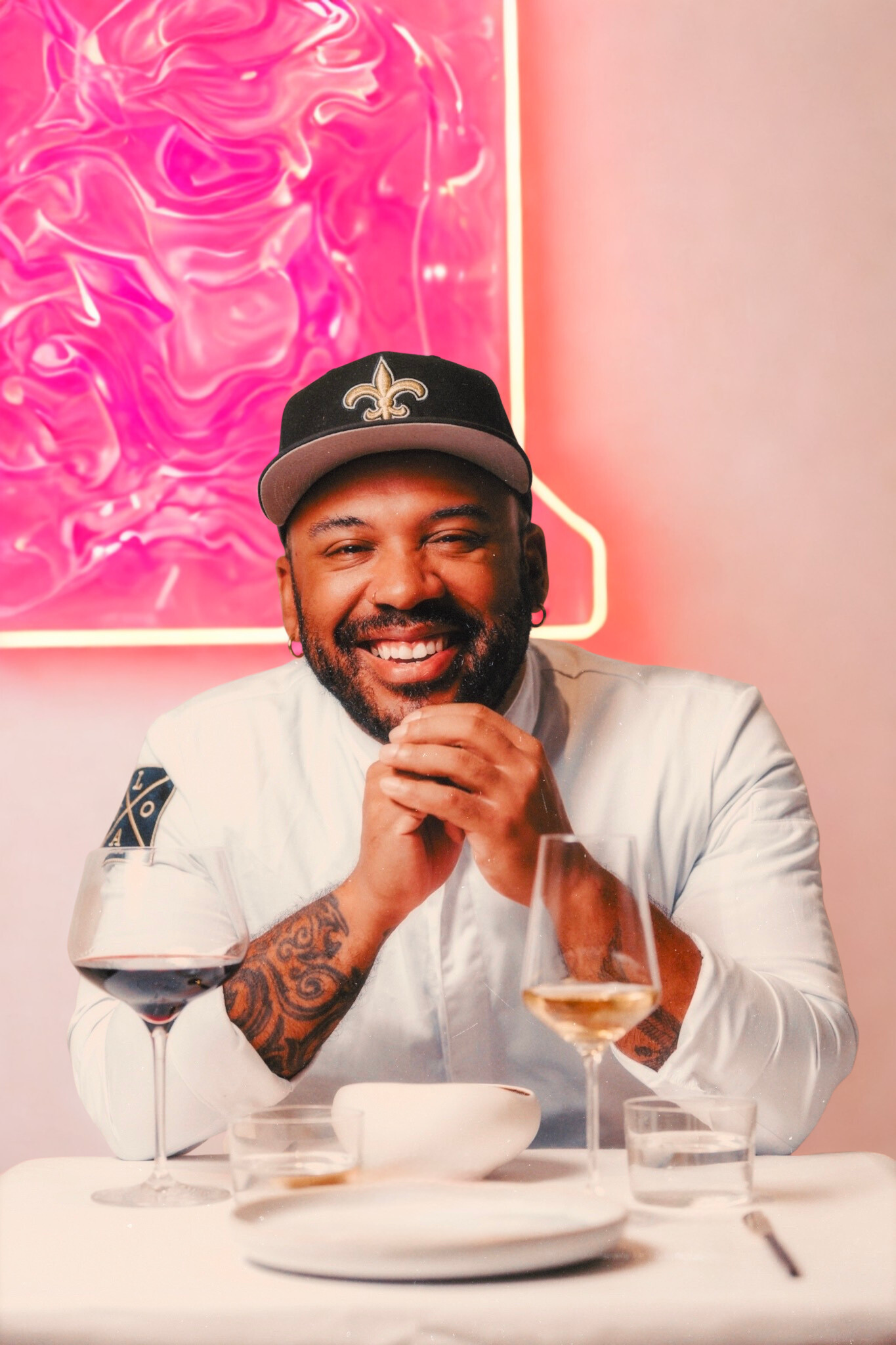 A smiling man with a beard and tattoos, wearing a black baseball cap with a fleur-de-lis symbol, sitting at a table with wine glasses and plates, in front of a pink neon art piece on a pink wall.