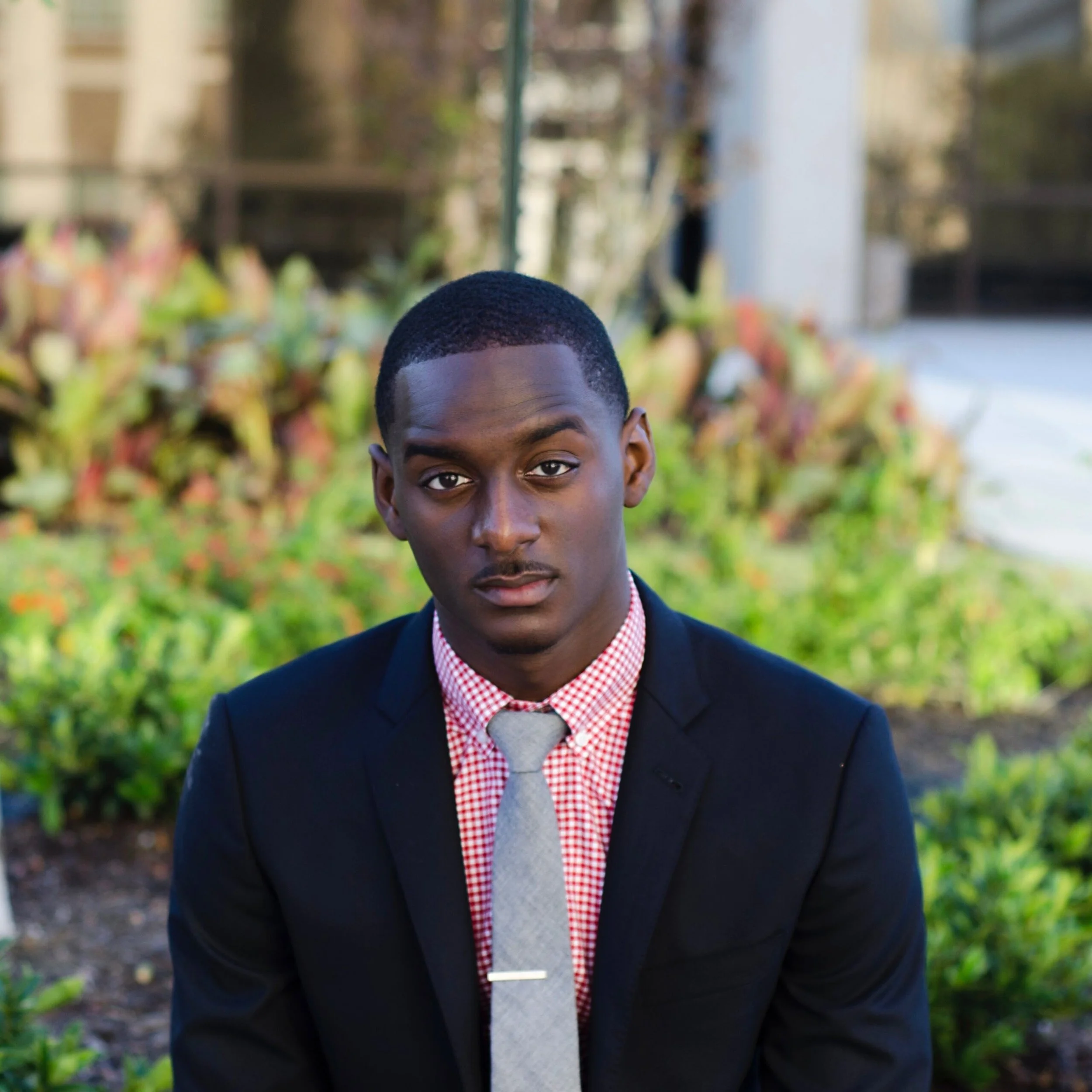 Chief Financial Officer, Charles, wearing a black suit jacket, red checkered shirt, gray tie, and a silver tie clip, sitting outdoors with greenery and a blurred background.