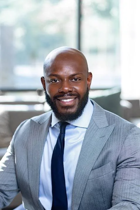 Portrait of executive director, Jevani Barron in a gray suit and blue tie, smiling, seated in an office with large windows in the background.