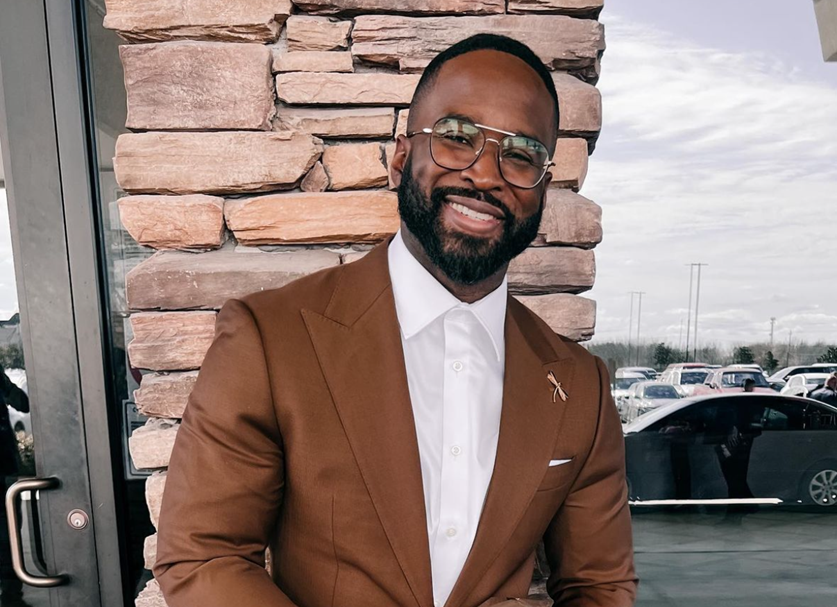 Image of Chief Clinical Officer, Tony, a man with a beard, glasses, and a big smile, dressed in a brown suit with a white shirt, standing outside near a stone wall and a parking lot.