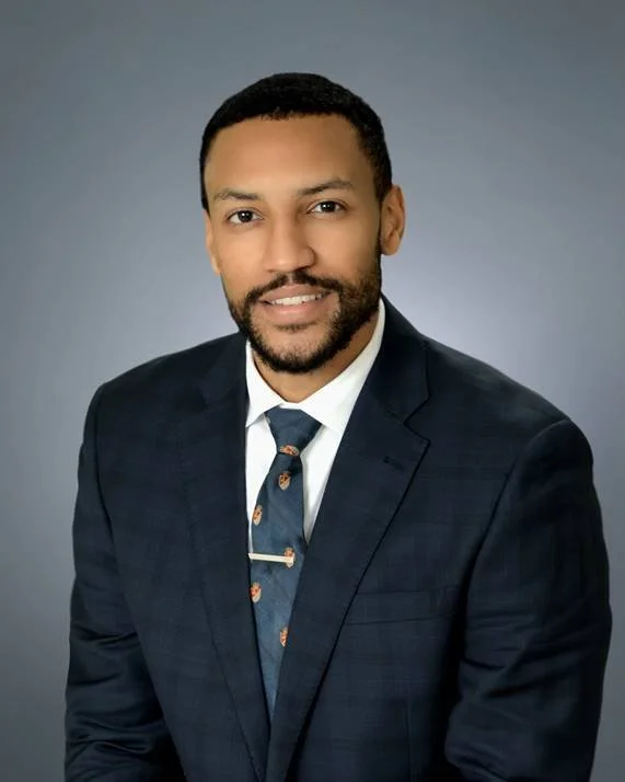 Professional portrait of Chief Legal Officer, Elliot, in a navy blazer, white shirt, and patterned tie, smiling against a gray background.