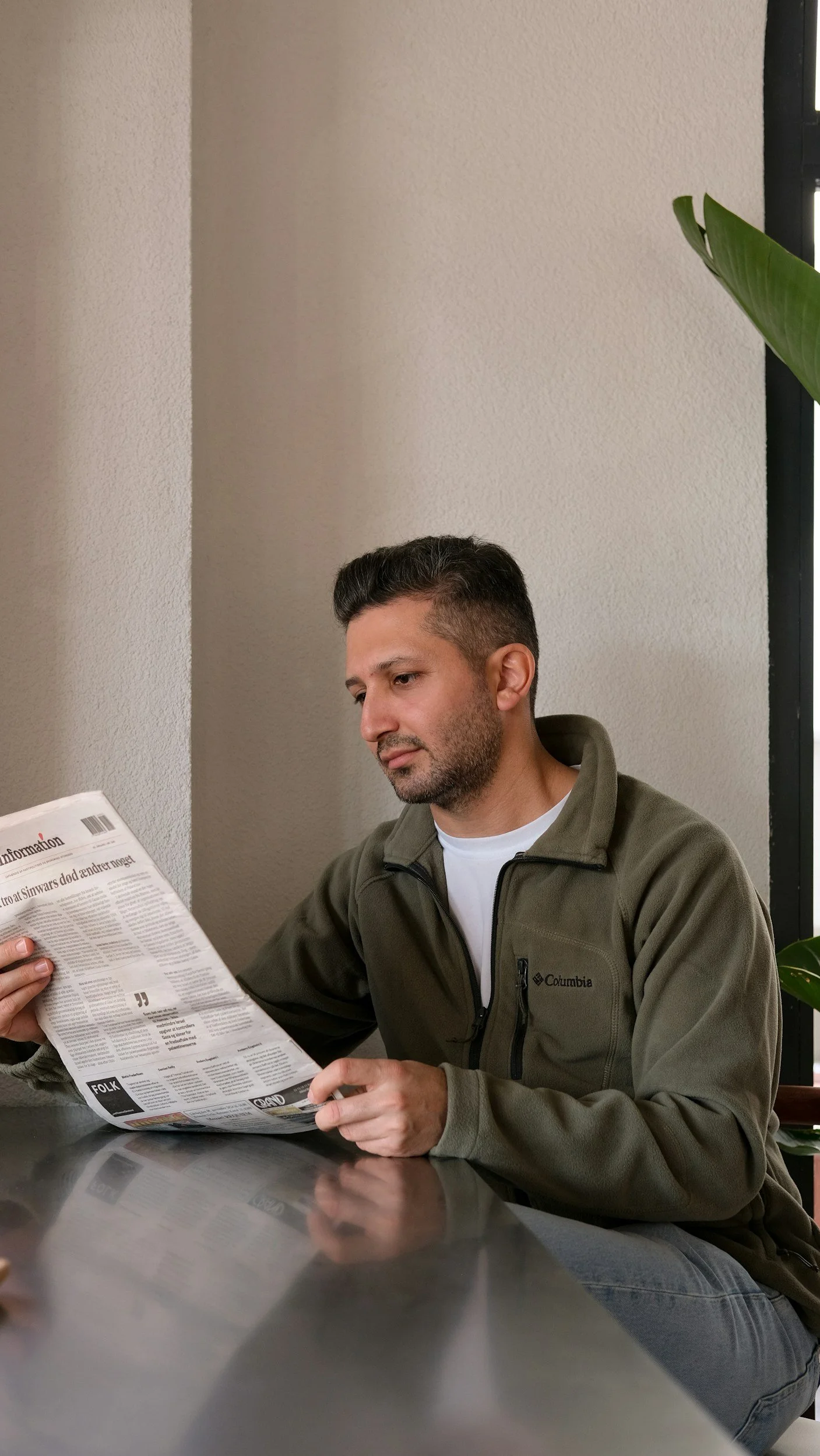 A man with short dark hair and a beard, wearing a gray Columbia fleece jacket, sitting at a reflective metal table, reading a newspaper in a room with beige walls and a large window with black framing, next to a green plant.