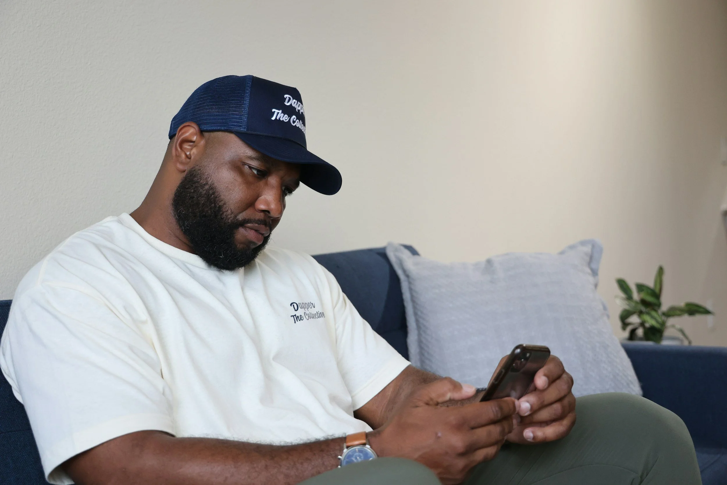 A man sitting on a blue sofa, looking at his smartphone. He is wearing a white T-shirt with 'Dupper The Collection' embroidered on it, a navy and mesh trucker cap with the same text, a wristwatch, and has a beard. There is a white pillow and a small green plant in the background.