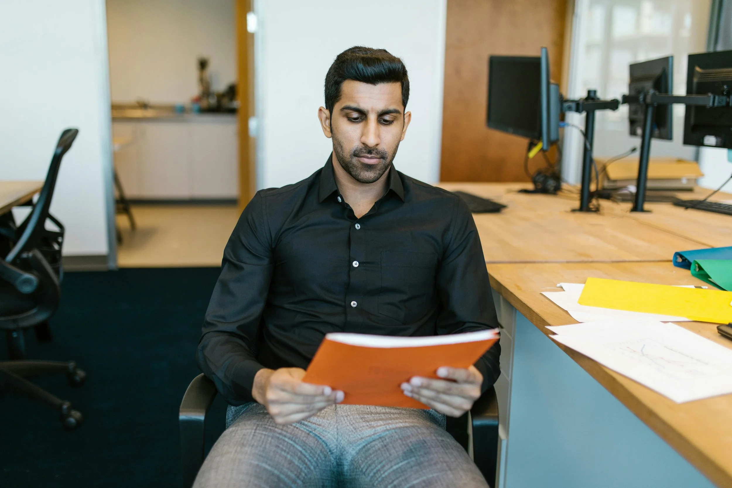 Man in black shirt sitting at desk, looking at orange folder in office