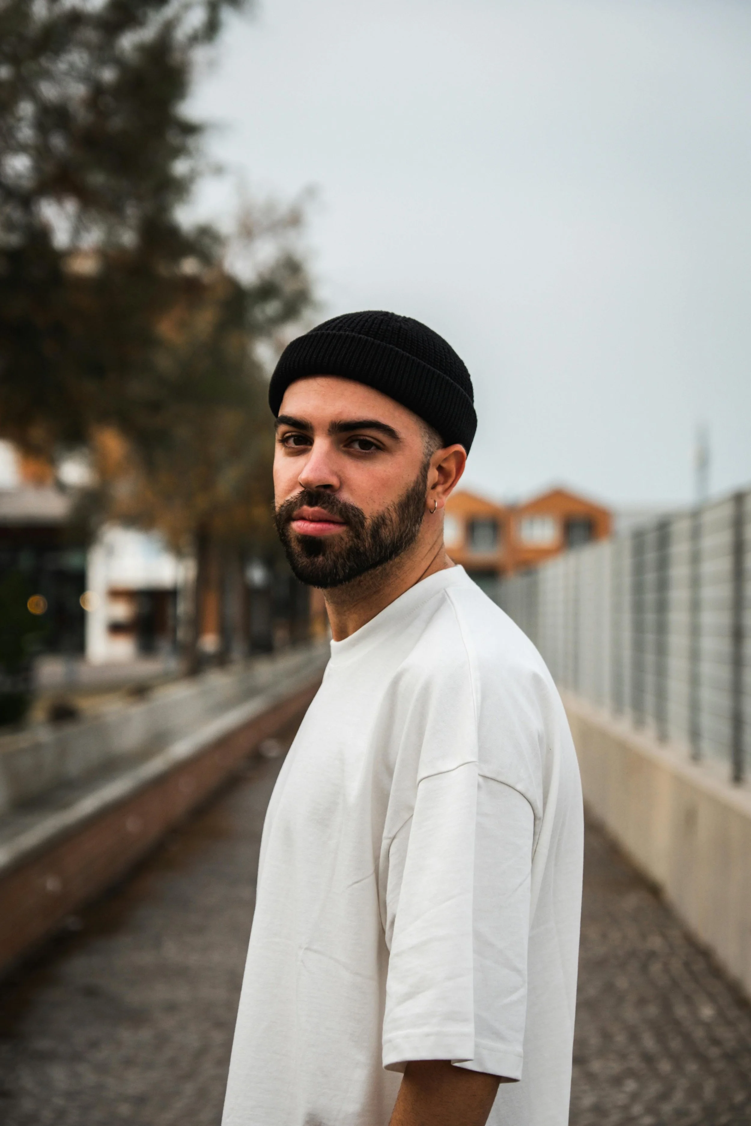 A young man with dark hair and a beard, wearing a black beanie and a loose-fitting white shirt, standing on train tracks outdoors on a cloudy day with blurred trees and buildings in the background.