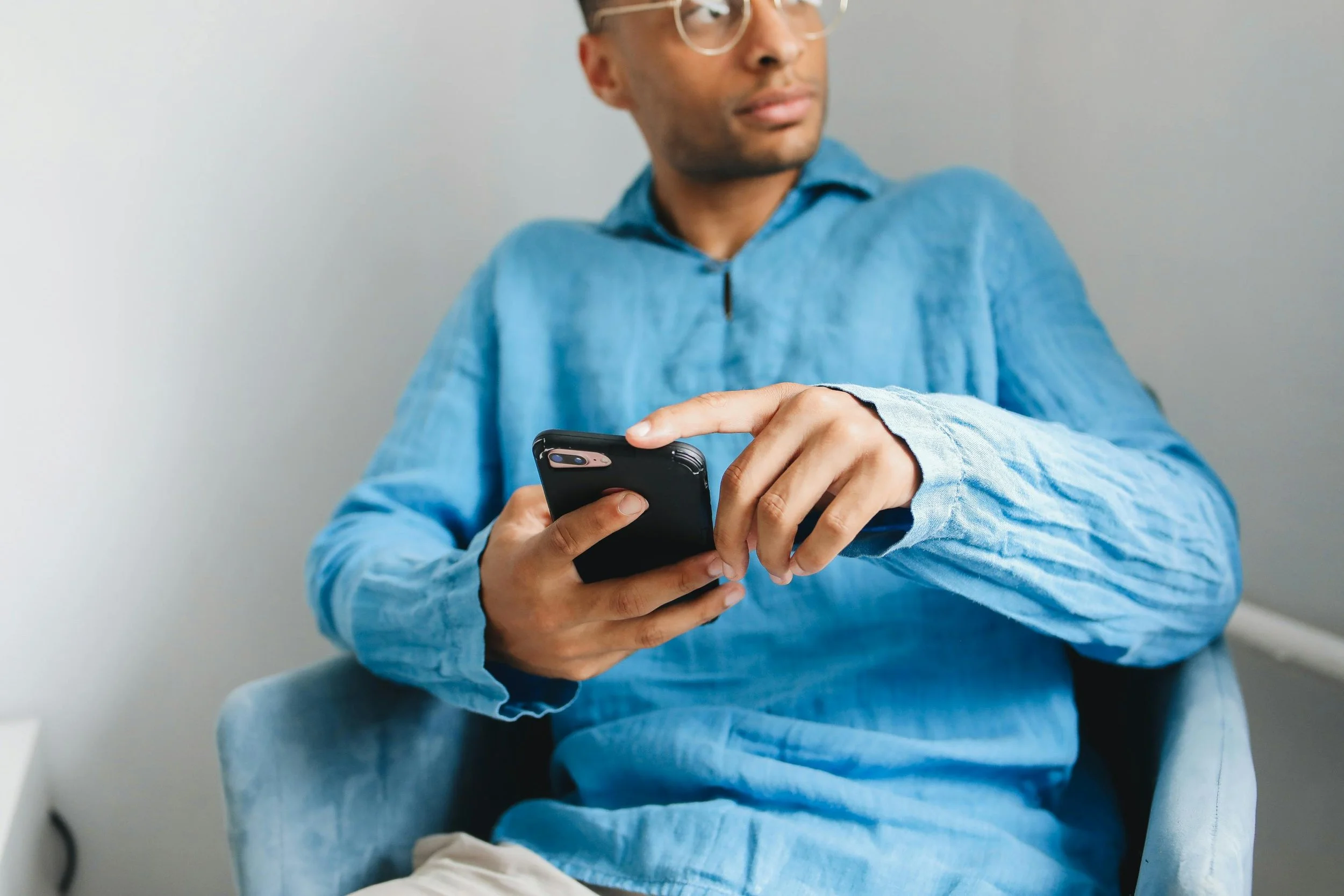 A man in a blue long sleeve shirt with glasses sitting in a chair, holding and using a smartphone.