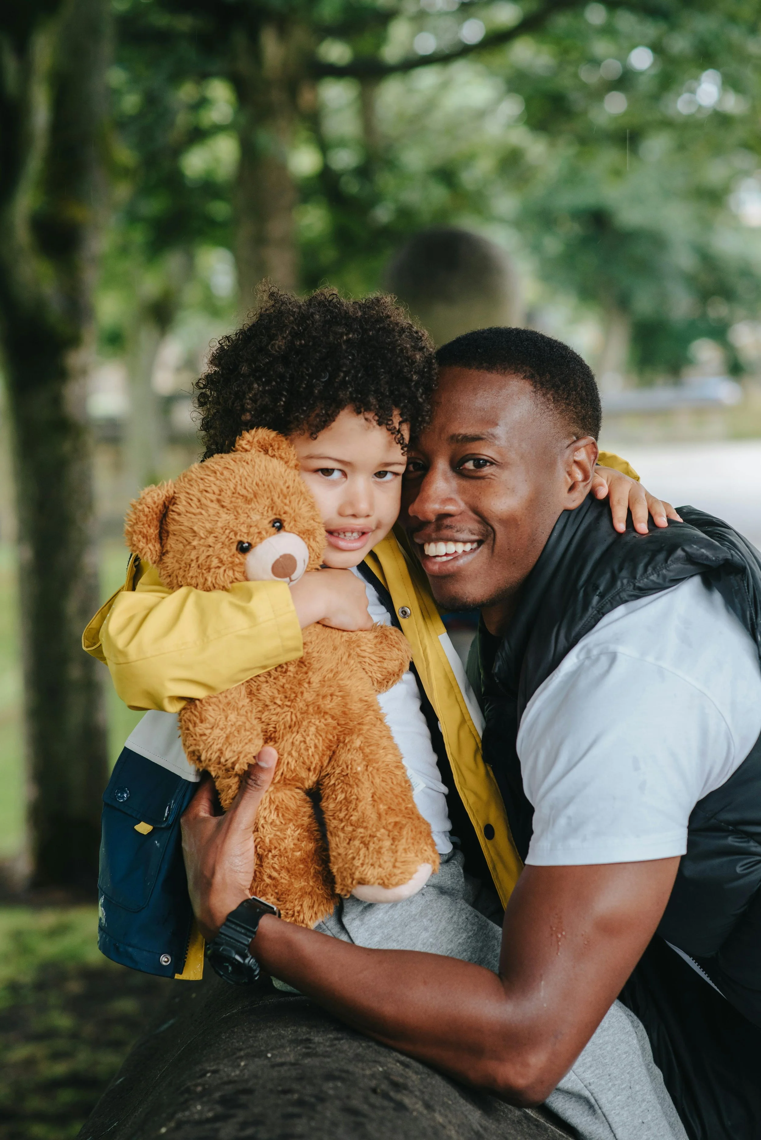 A father with short hair and a beard, smiling, holds his young son with curly hair, who is hugging a teddy bear, outdoors in a park with trees in the background.