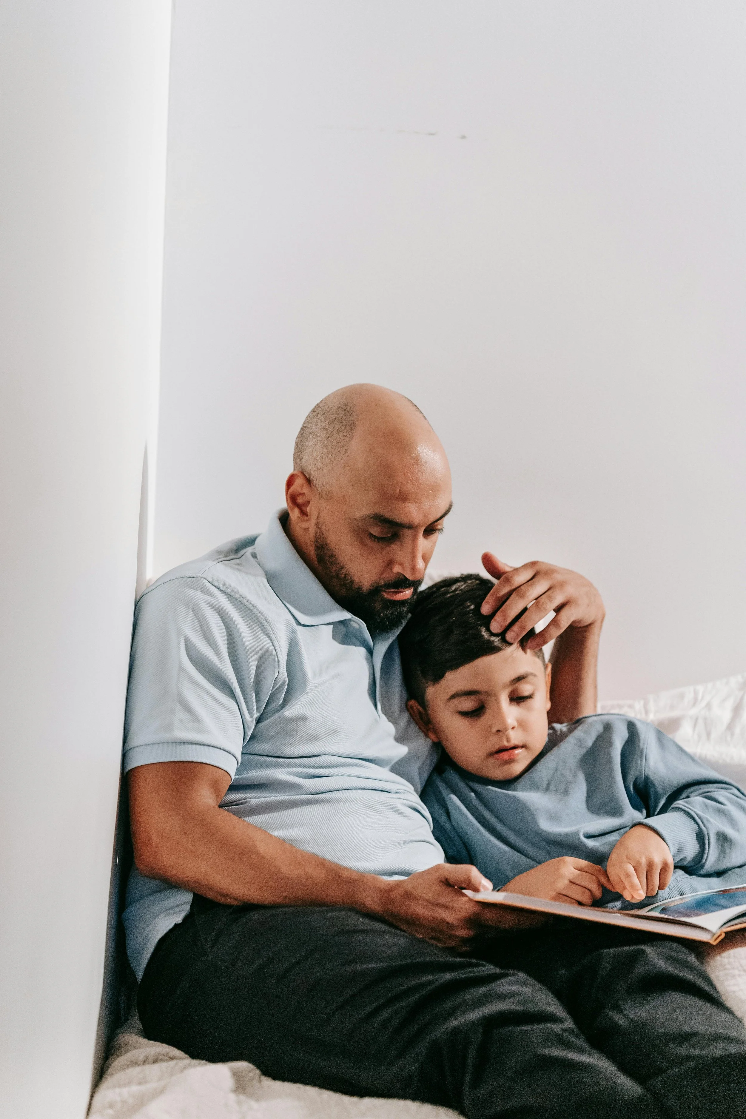 A man and a young boy sitting together on a bed, reading a book. The man has a shaved head and a beard, wearing a light blue polo shirt. The boy has dark hair, wearing a blue sweatshirt. The man is resting his head on the boy's, and they both appear focused on the book.