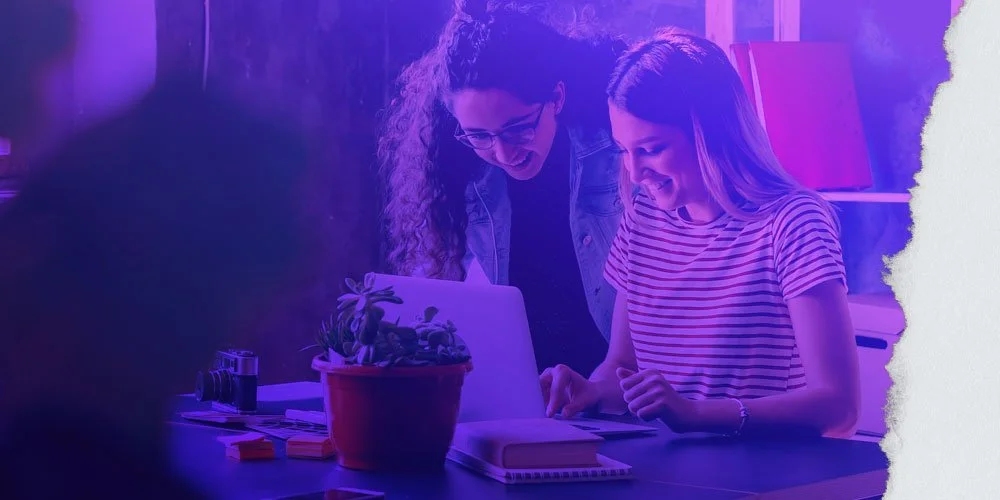 Two young women are smiling and looking at a laptop on a desk, with books and a potted plant nearby, in a room with purple lighting.