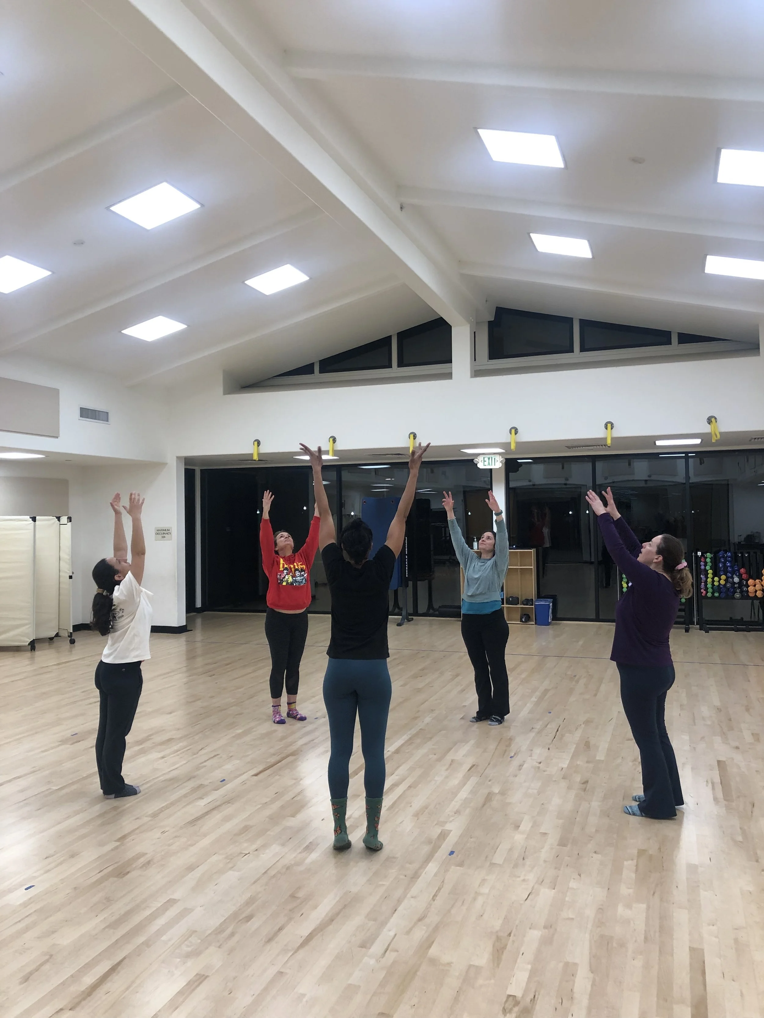 Five women in workout clothes standing in a circle on a wooden floor in a spacious, well-lit room, raising their arms during a fitness class or exercise session.