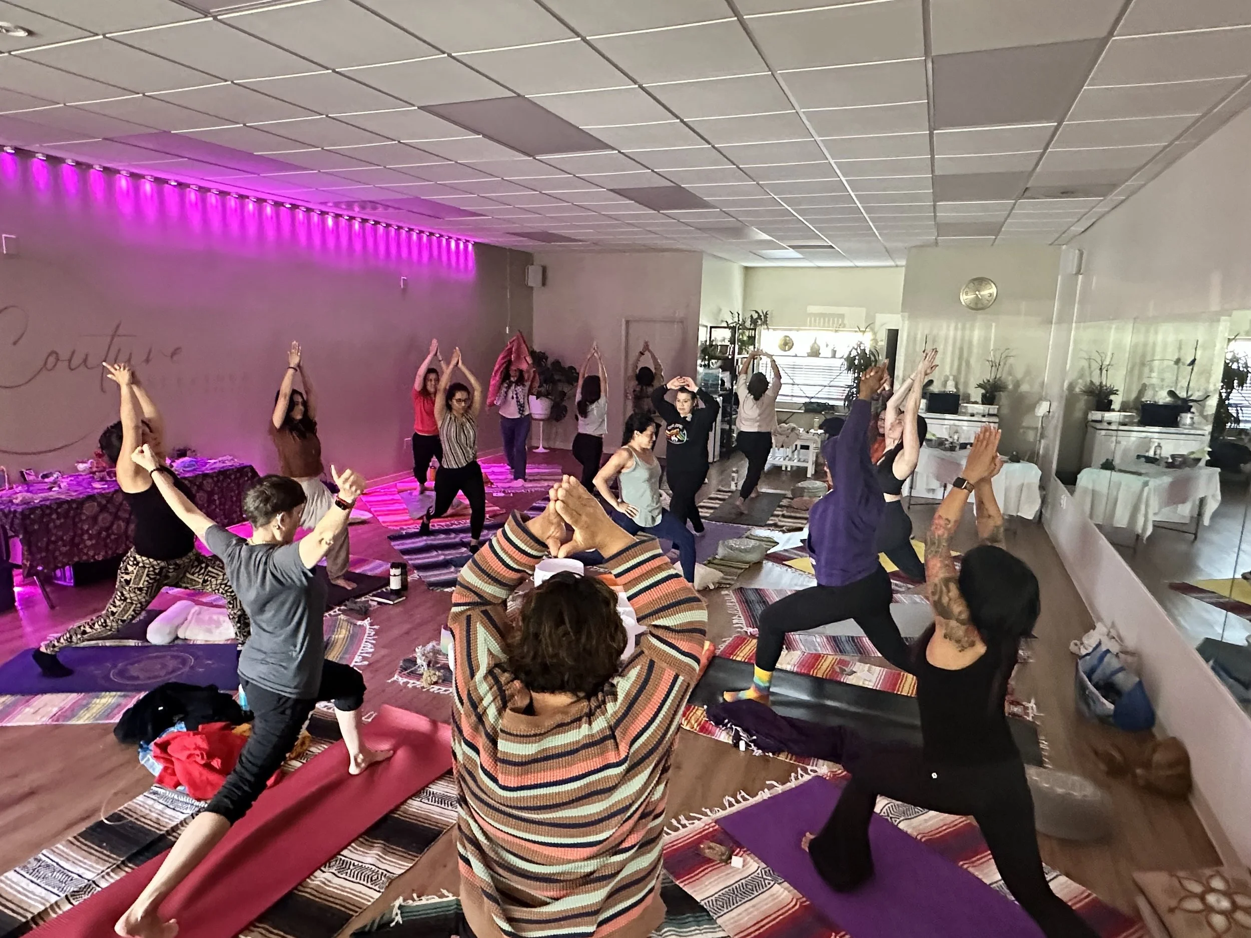 People participating in a yoga class in a room with mirrors and potted plants, practicing poses on mats.