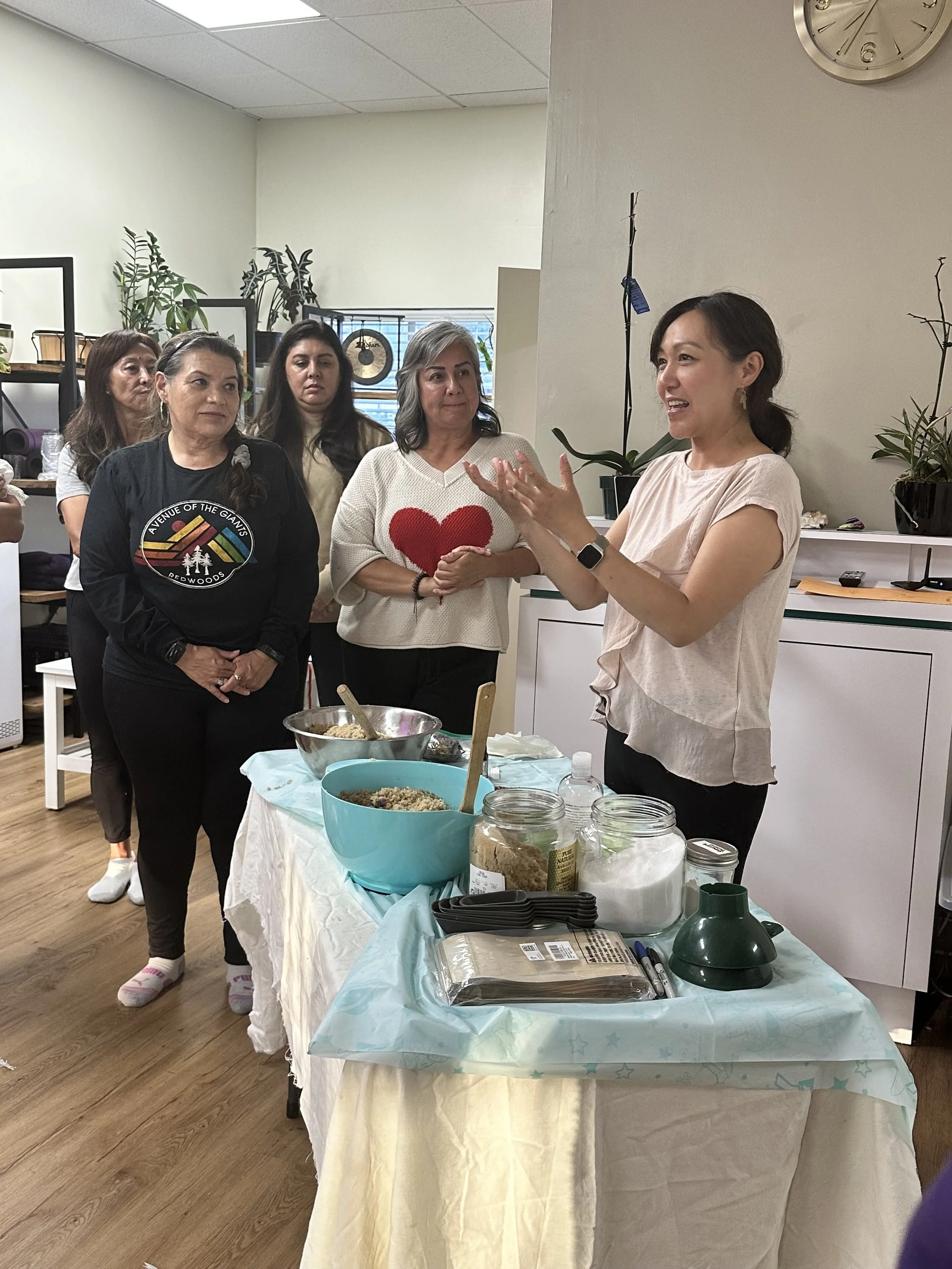 A woman giving a cooking demonstration to a group of women in a kitchen setting with ingredients and utensils on the table.