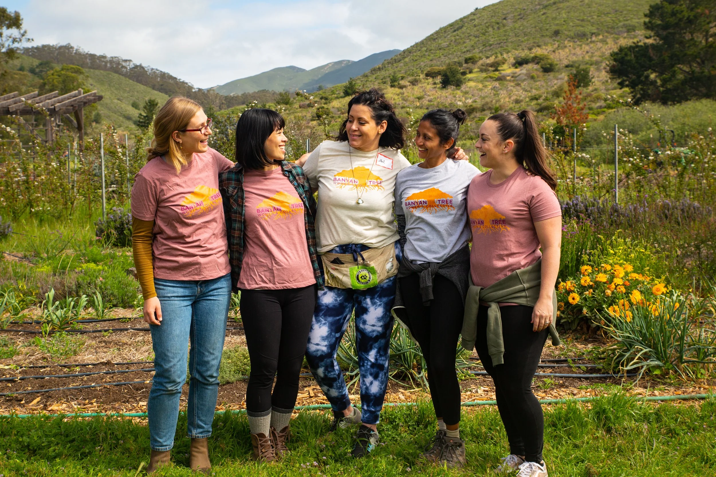 Five women standing arm-in-arm outdoors, smiling, in a lush garden with mountains in the background, wearing t-shirts with the logo 'Banyan Tree Women's Collective'.