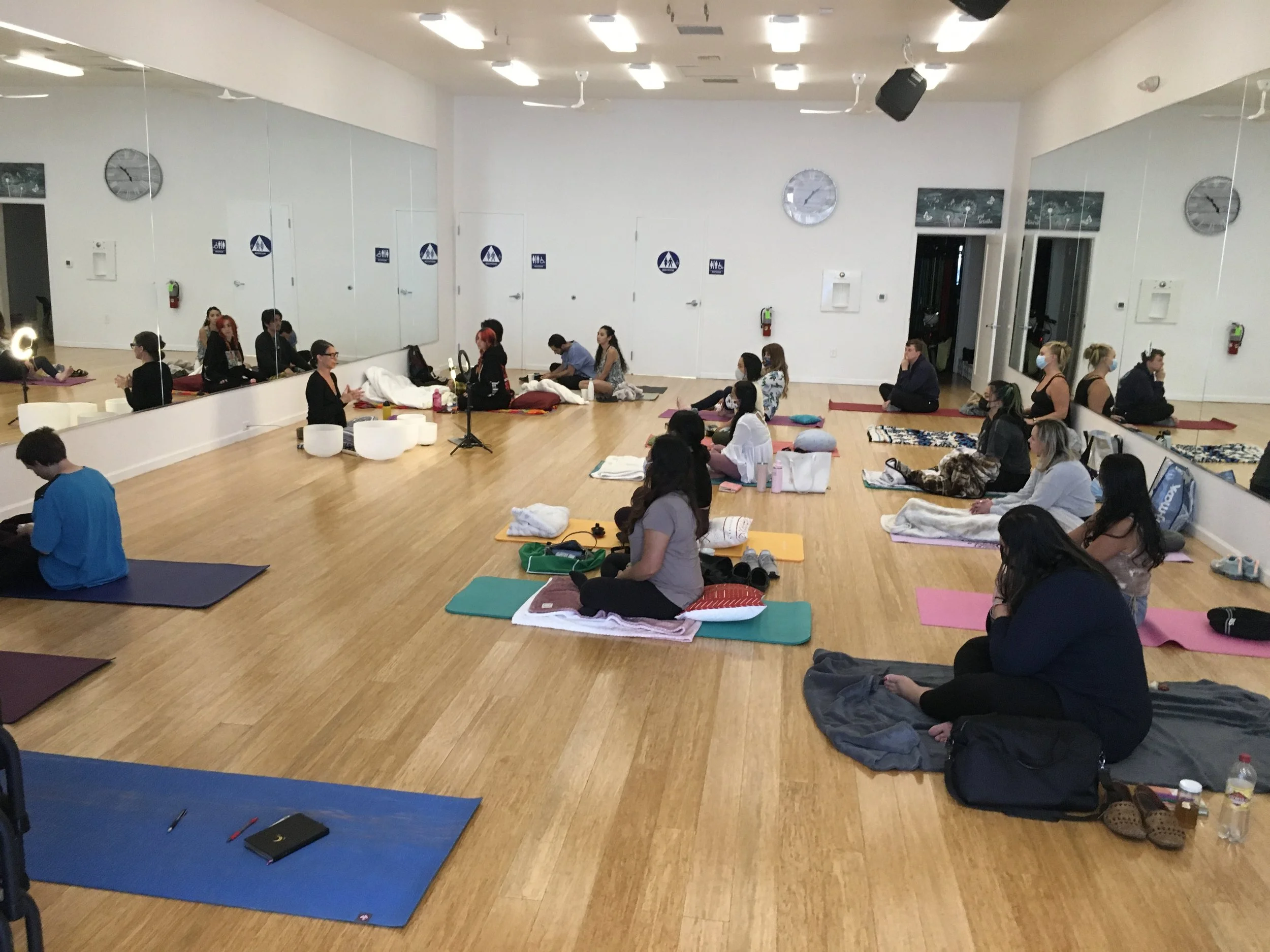 People practicing yoga or meditation in a dance studio with wooden floors, large mirrors, and wall clocks.