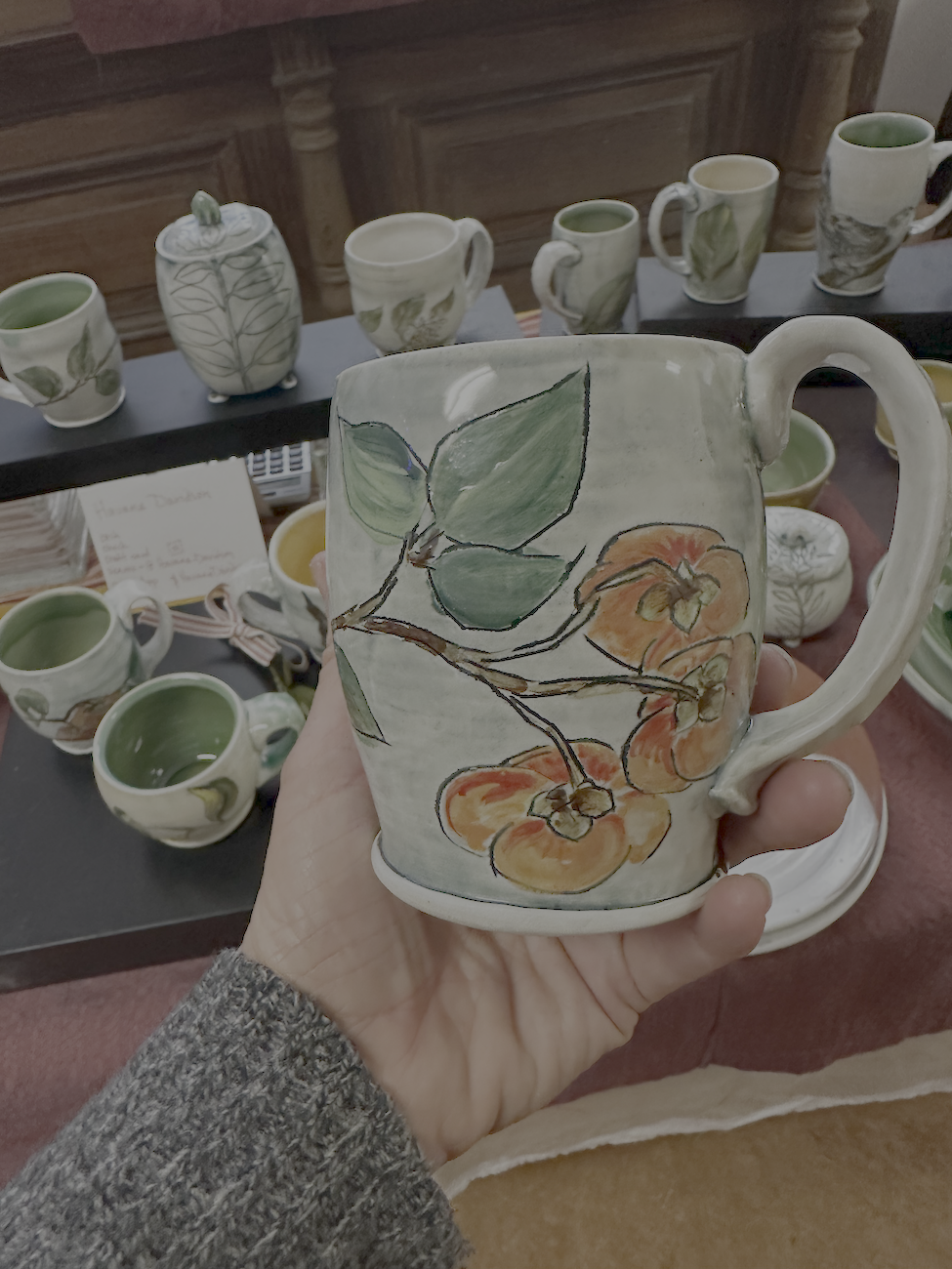 Ceramic mug decorated with painted persimmons and green leaves, with a display of similar mugs on shelves in the background.