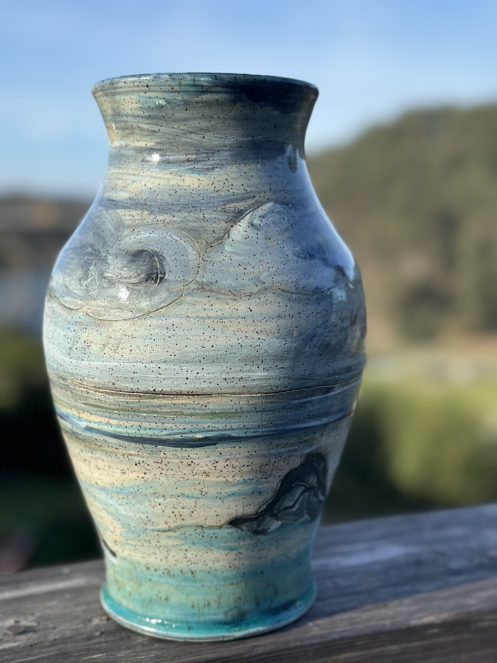 Close-up of a ceramic vase with swirling blue, gray, and cream patterns, set outdoors on a wooden surface with a blurred landscape in the background.