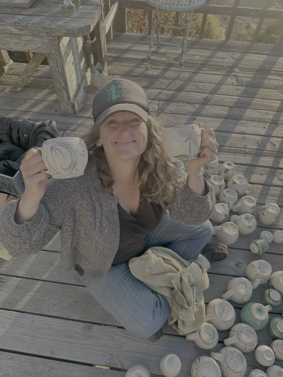A woman sitting cross-legged on a wooden deck, holding two decorated ceramic mugs, with numerous more mugs scattered around her, sun shining on her face, and a weathered wooden table nearby.