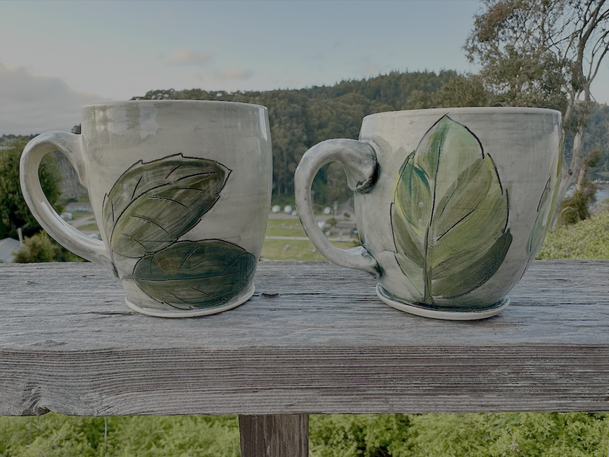 Two ceramic mugs with green leaf designs on a wooden surface outdoors, with trees and hills in the background.