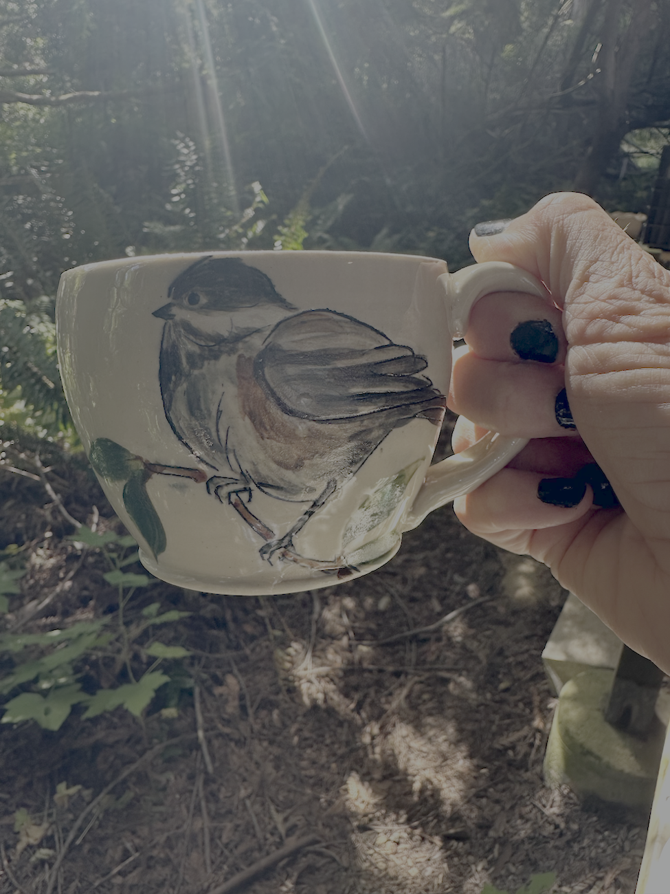 A hand holding a ceramic mug with a painted bird, outdoors with sunlight shining through trees.