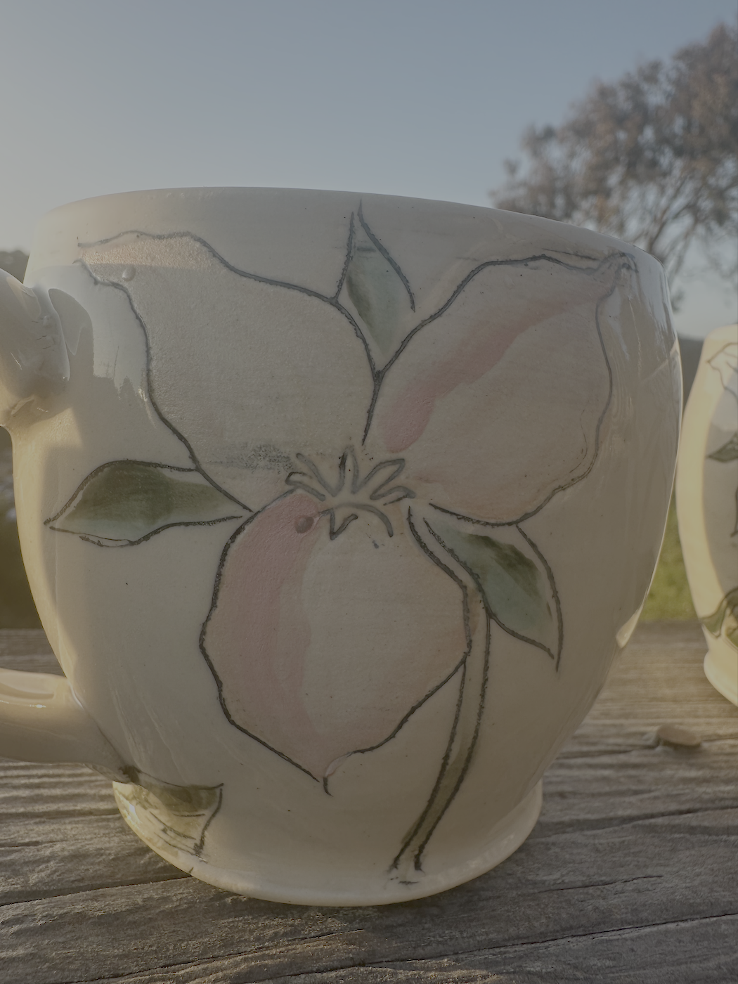 Close-up of a ceramic mug with a painted flower design, featuring pink and peach blossoms and green leaves, placed on a weathered wooden surface outdoors in bright sunlight.