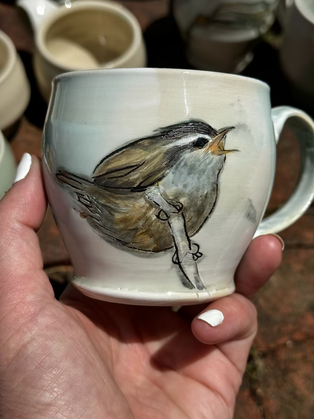 A ceramic mug with a painted illustration of a bird, held in a person's hand, with additional ceramic mugs in the background.