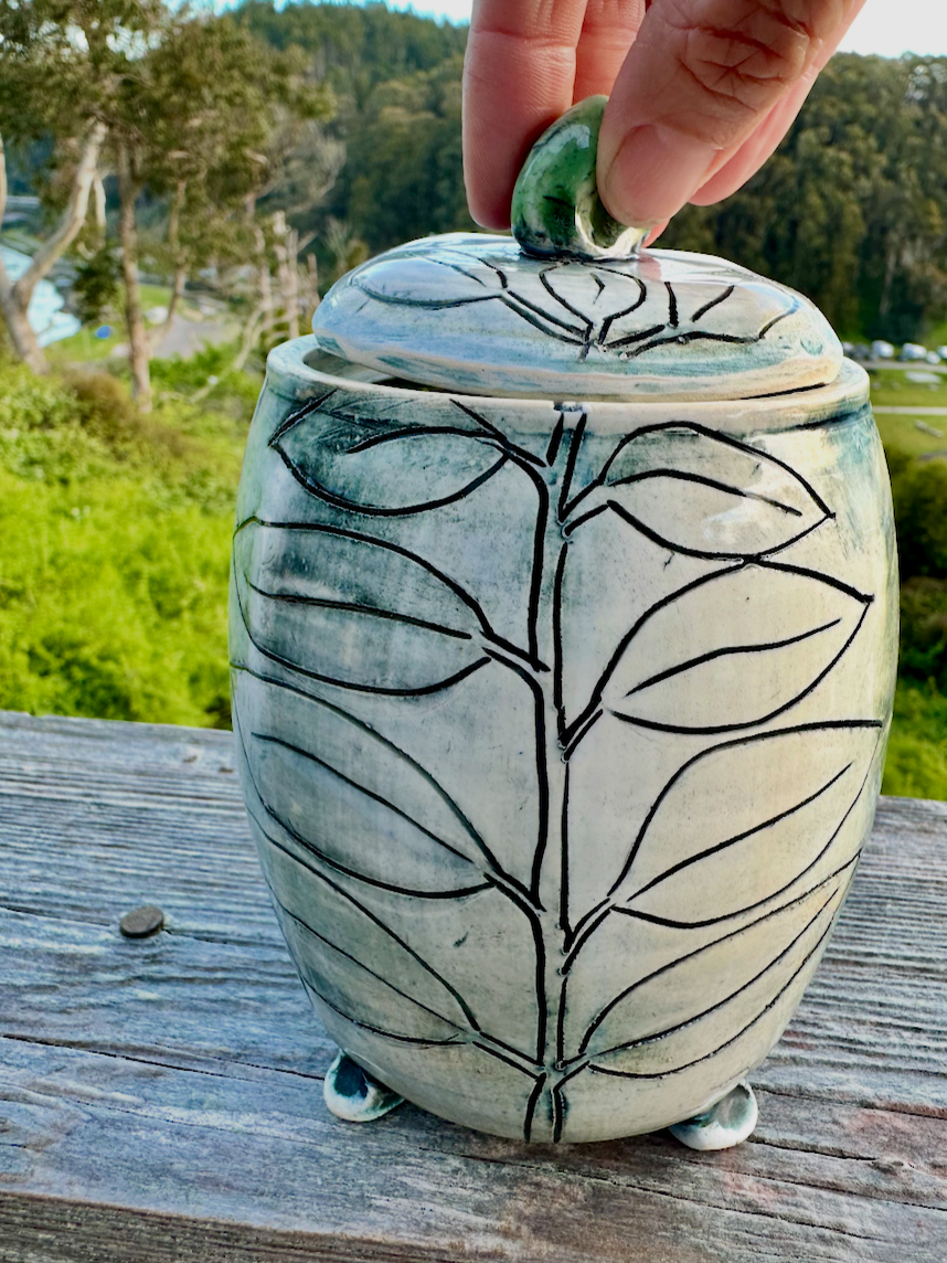 A ceramic jar with a lid being lifted by a person's hand. The jar has a leaf pattern design on its surface, set outdoors on a weathered wooden surface with greenery and trees in the background.