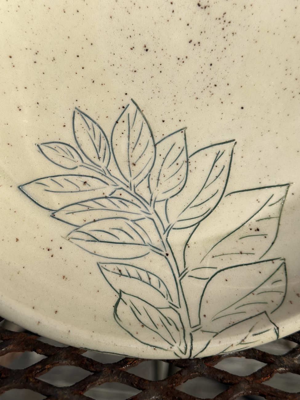 A ceramic or porcelain surface with a line drawing of a plant, featuring multiple leaves, and a speckled pattern on a beige background.