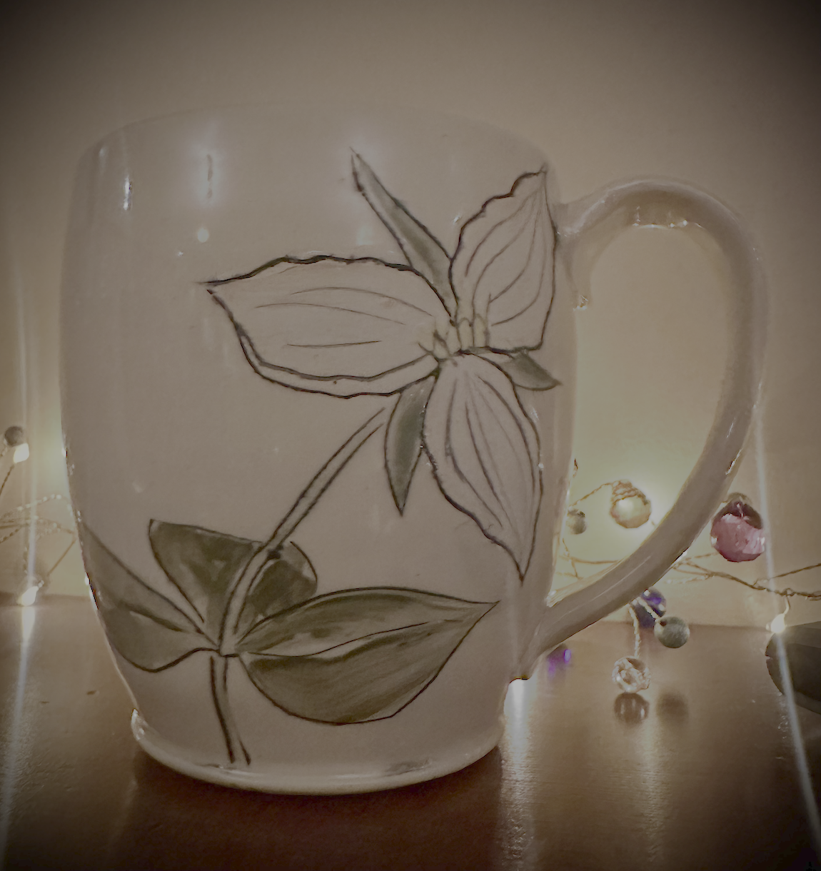 A ceramic mug with a floral design featuring a large flower and leaves, illuminated by warm lighting, with small decorative beads or ornaments around it.