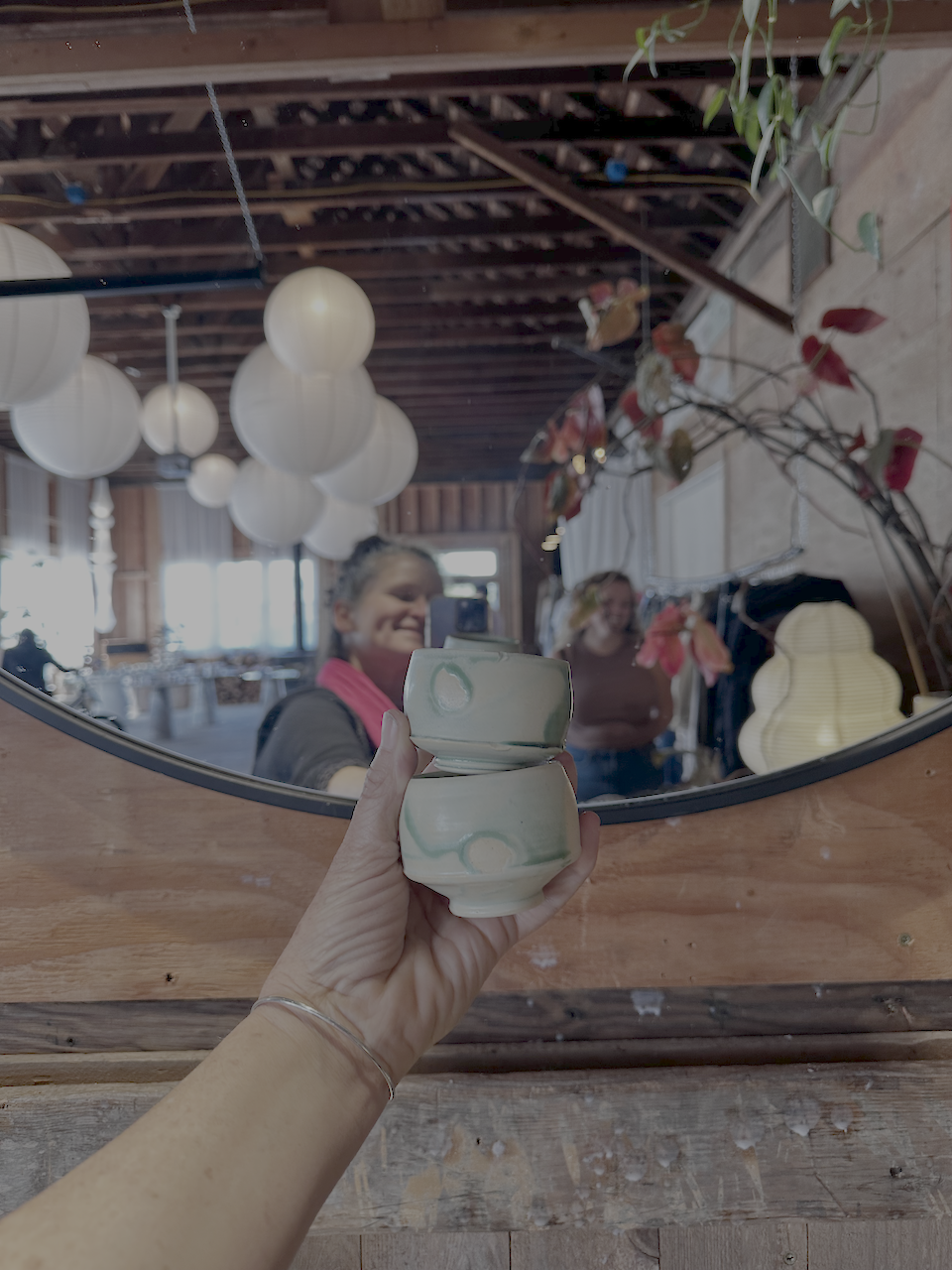 A hand holding two small ceramic cups in front of a mirror. The reflection shows two smiling women inside a room decorated with paper lanterns and hanging lamps.