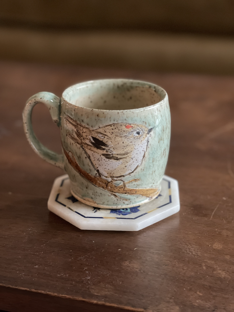 A ceramic mug with a bird design resting on a small white and blue tile coaster on a wooden surface.