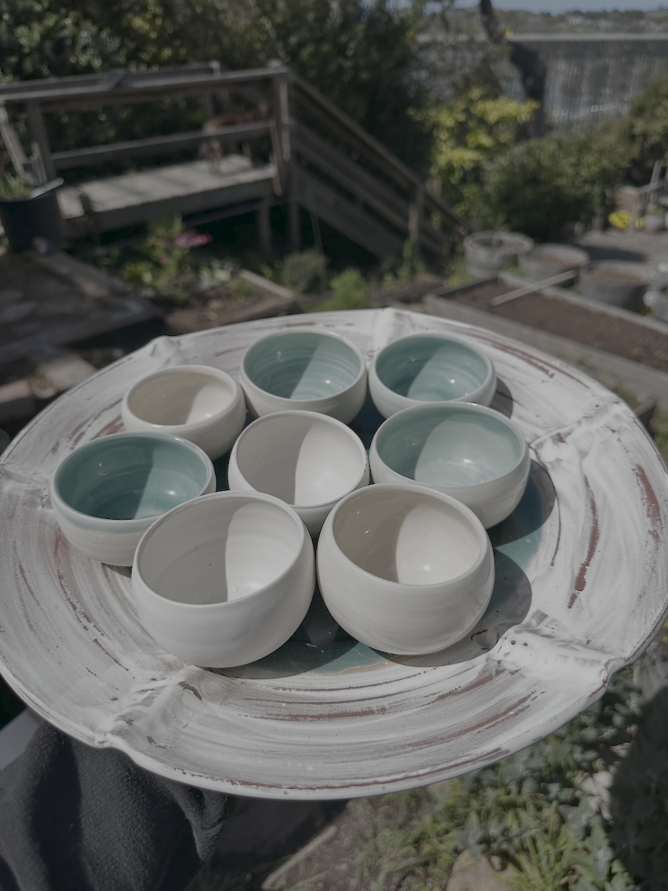 Hand holding a white ceramic plate with several small, round bowls that are white on the outside and glazed in shades of green and blue on the inside, outdoors in a garden.