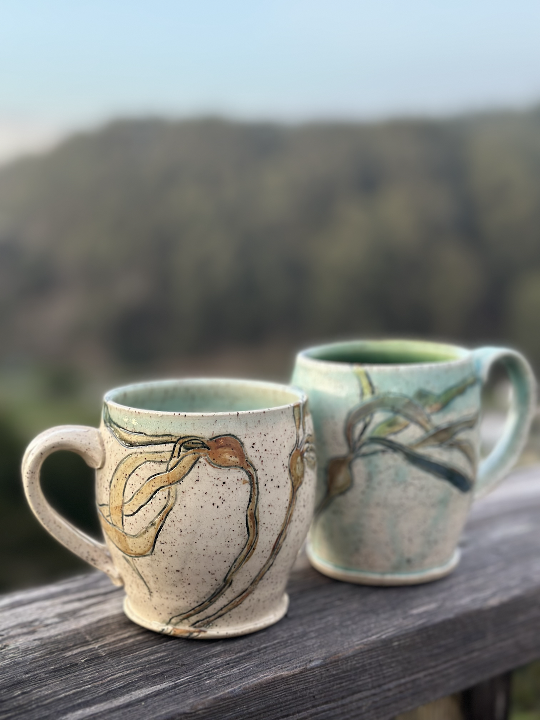 Two ceramic mugs with painted abstract designs on a weathered wooden surface, with a blurred natural landscape background.