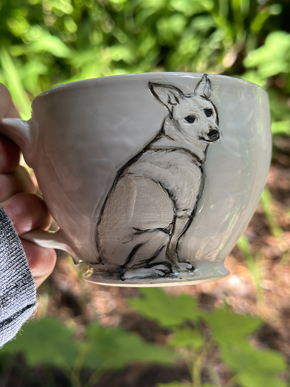 A ceramic mug with a painted image of a sitting dog, set against a blurry green outdoor background.