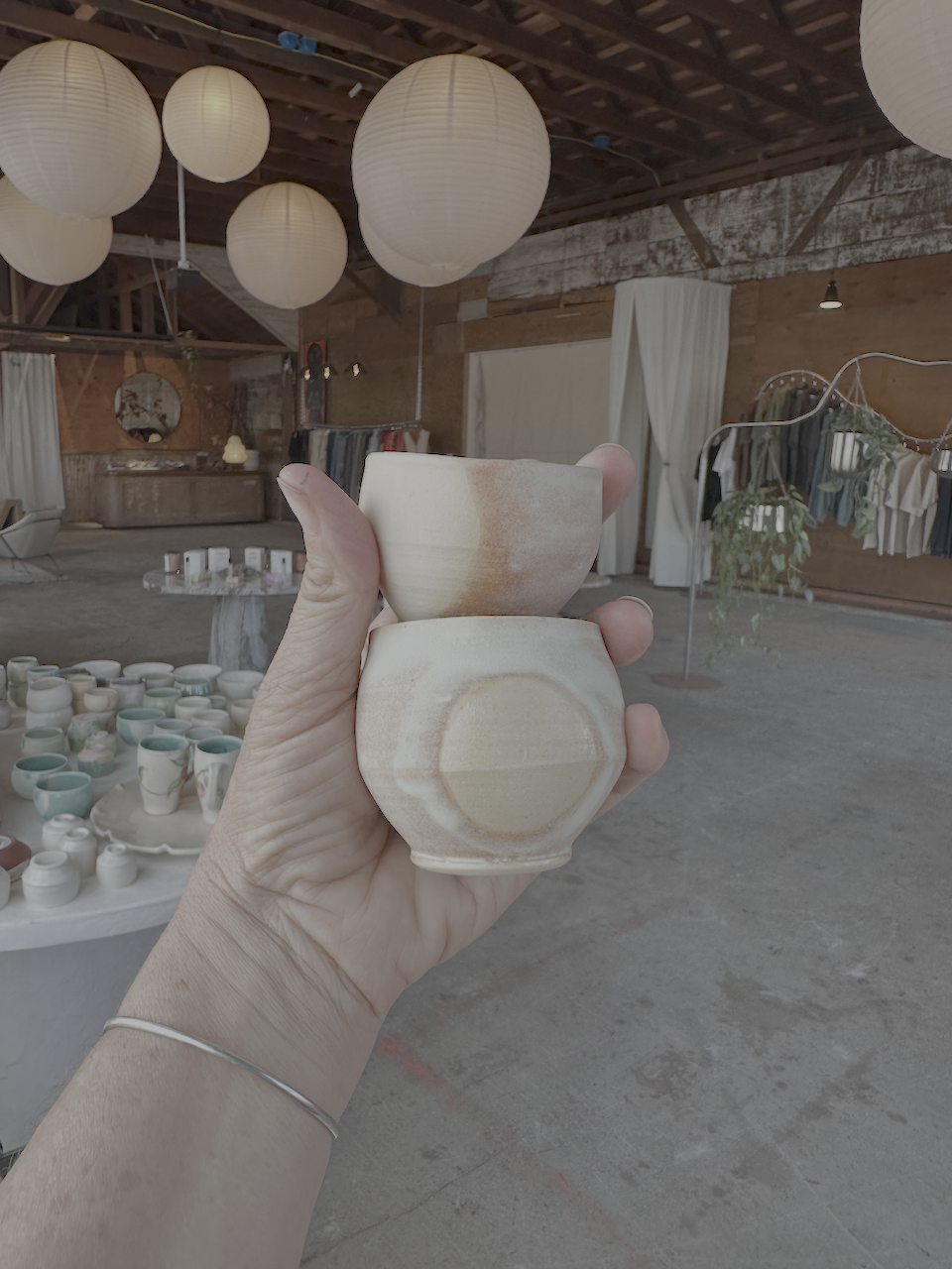 Person holding two stacked ceramic bowls in an indoor craft or market space with hanging paper lanterns and display tables of pottery and clothing.