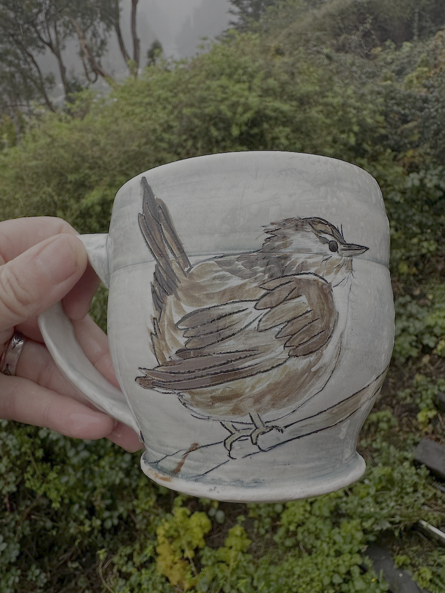 A hand holding a ceramic mug painted with a brown and gray bird outdoors with green bushes and trees in the background.