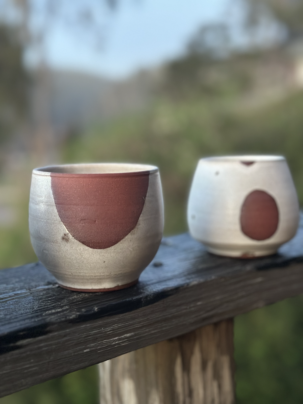 Two ceramic cups with red semi-circular designs on a wooden railing, outdoors with blurred trees and sky in the background.