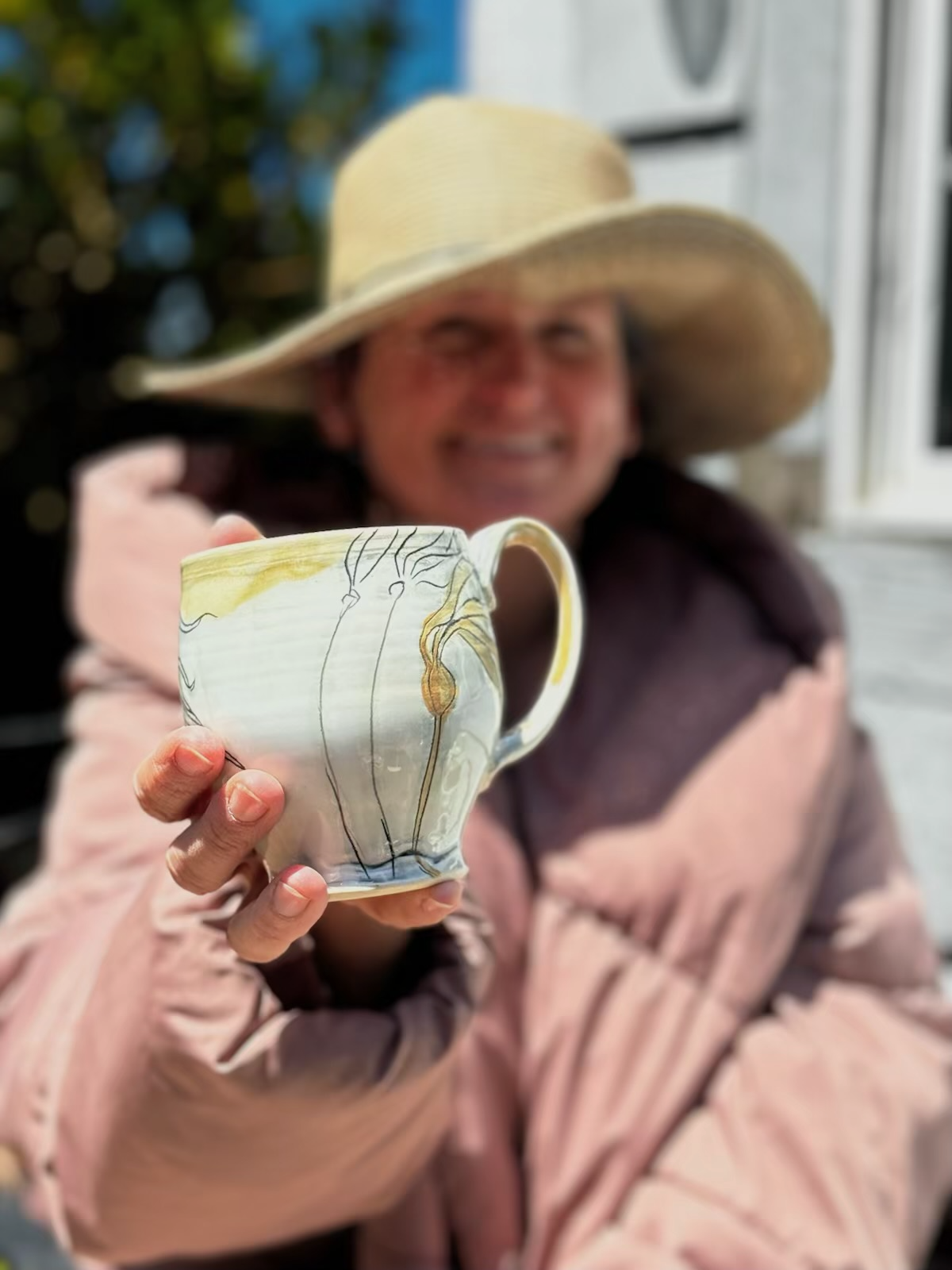 Person wearing a large straw hat holding a white ceramic mug with abstract yellow, black, and gray patterns, smiling in the background.