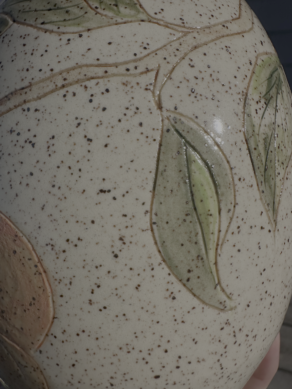 Close-up of a ceramic object with a speckled cream and brown glaze, featuring hand-painted green leaves and brown outlines.