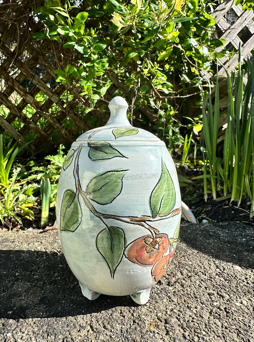 Decorative ceramic jar with a lid, painted with a design of green leaves and brown fruit on a garden surface with green plants and a wooden lattice in the background.