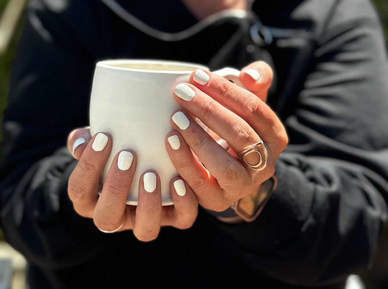 A person holding a white mug with both hands, showing white painted nails and wearing a ring on their right index finger, with a dark jacket.