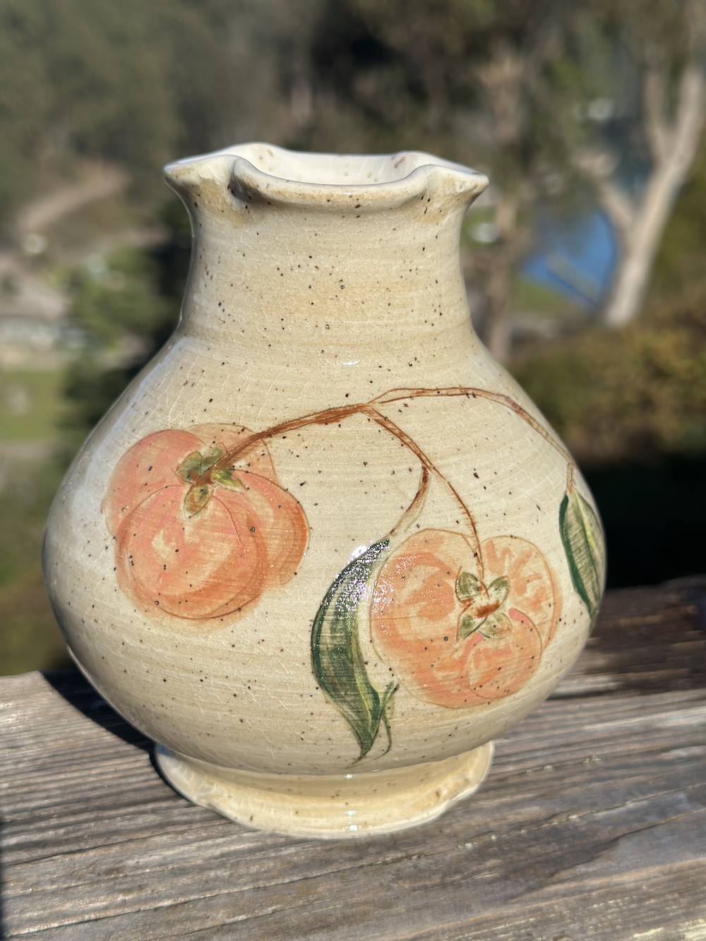 Decorative ceramic vase with painted orange-yellow cherries and green leaves, placed on a wooden surface outdoors.