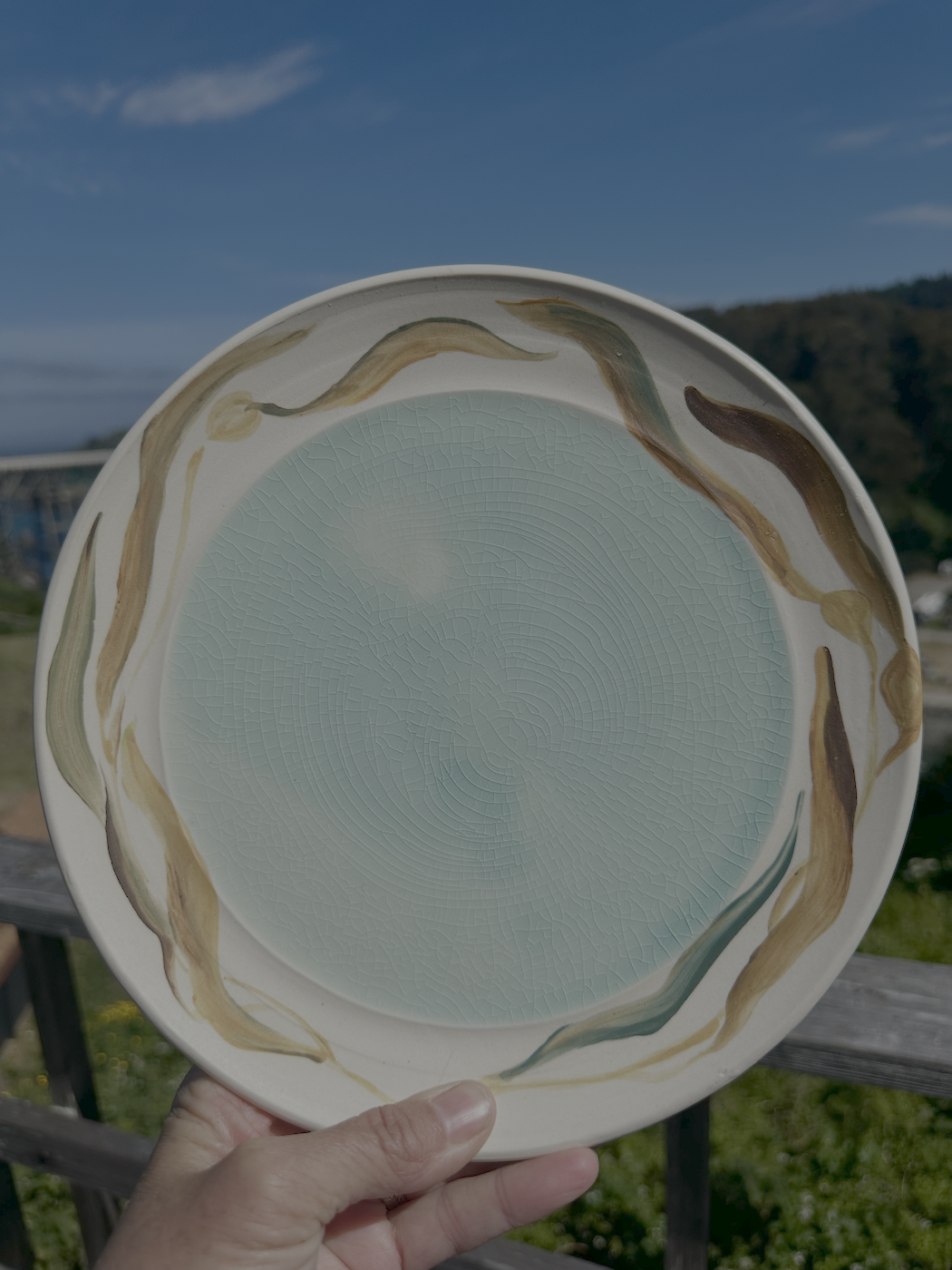 A hand holding a ceramic plate with a crackle glaze design, featuring blue-green color with brown, green, and tan decorative swirls around the rim, outdoors with a blue sky and mountains in the background.