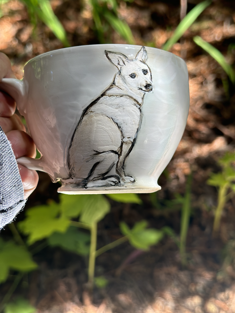 A white mug with a painted illustration of a sitting dog looking back over its shoulder, held by a person's hand outdoors with green plants and soil in the background.