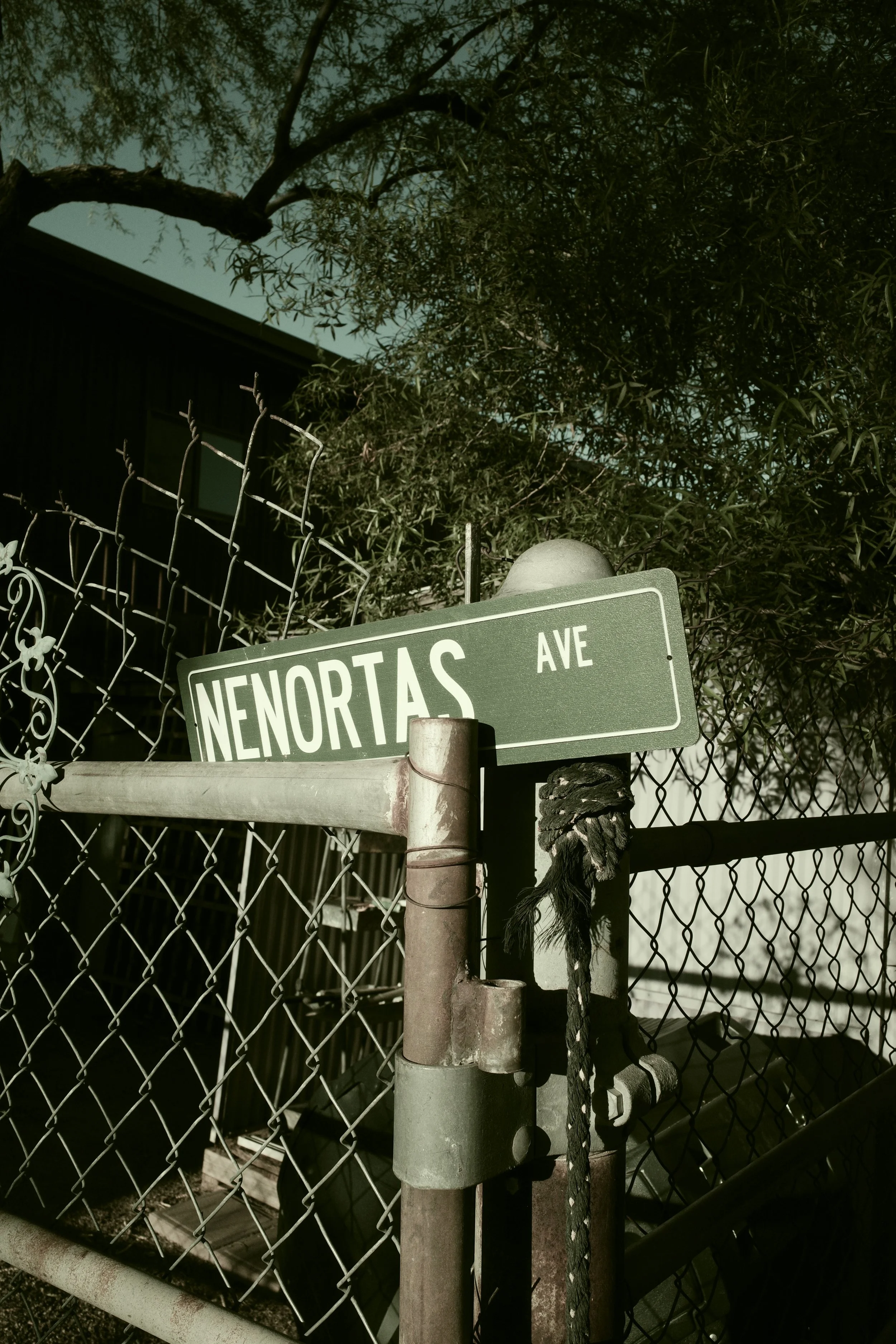 Street sign at Nenortas Avenue attached to a weathered metal pole, with a chain-link fence and greenery in the background.