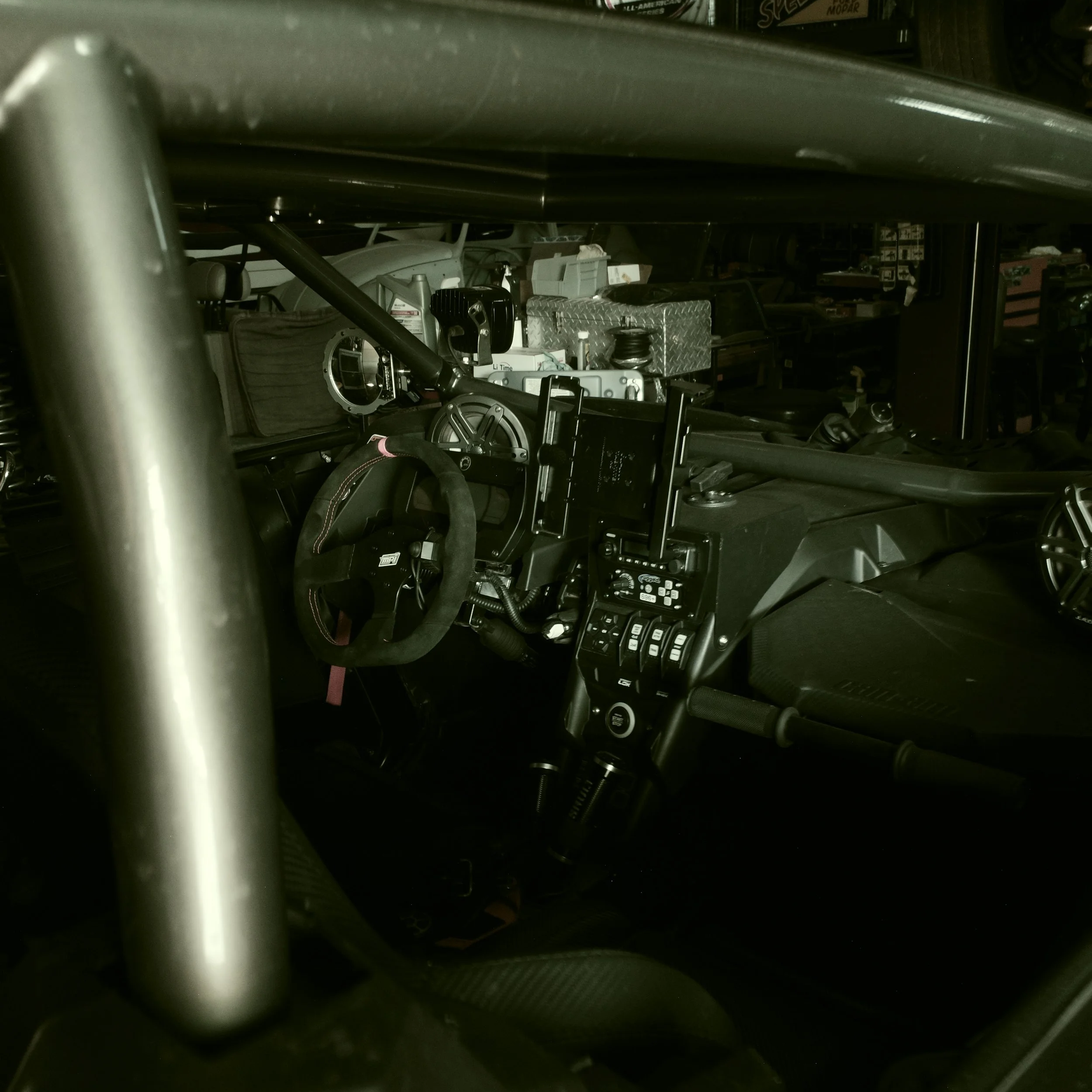 Close-up of the interior of a race car, showing the steering wheel, dashboard, and various control panels, with a storage area filled with equipment in the background.