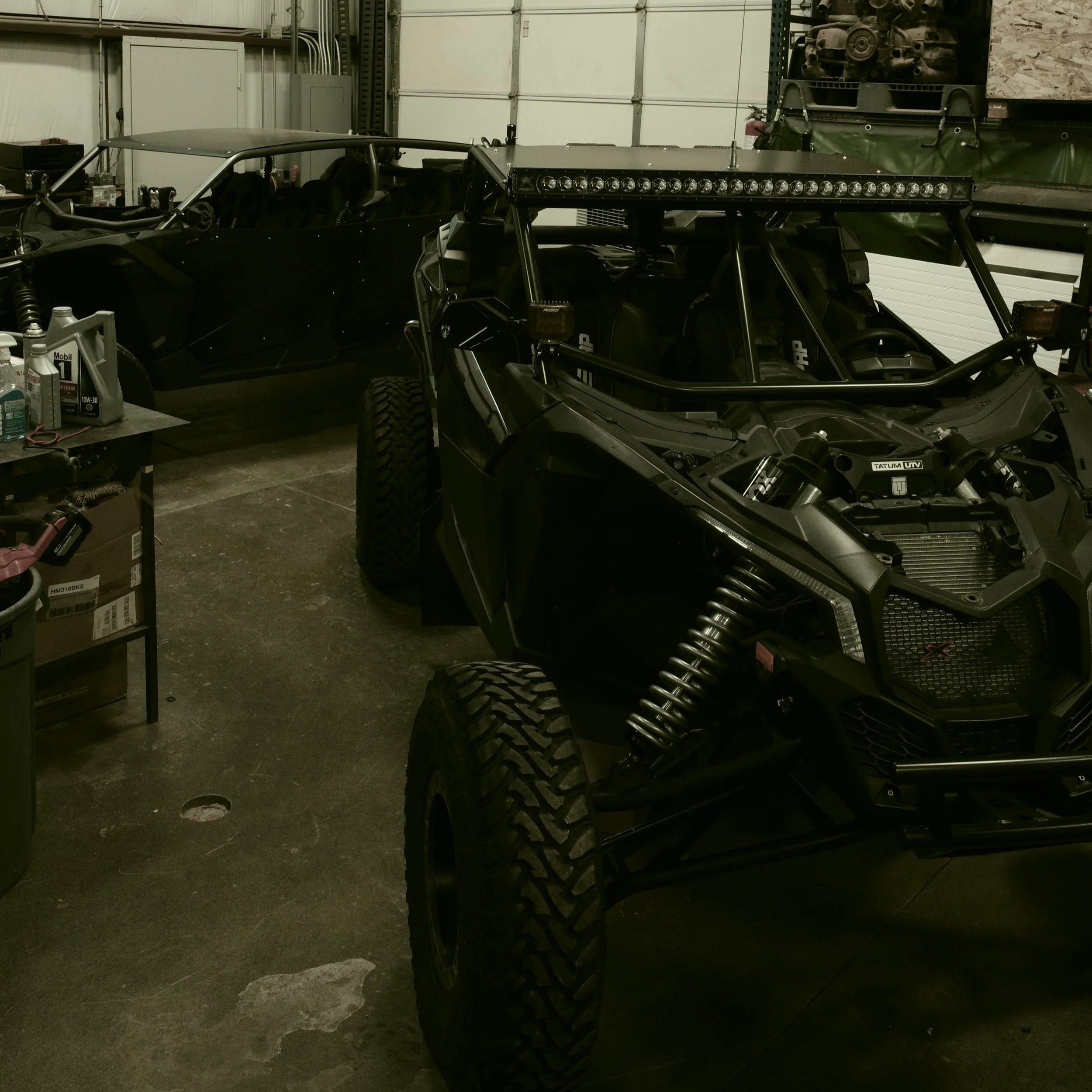 A black off-road vehicle with large tires, suspension, and a roll cage parked inside a garage near another similar vehicle.