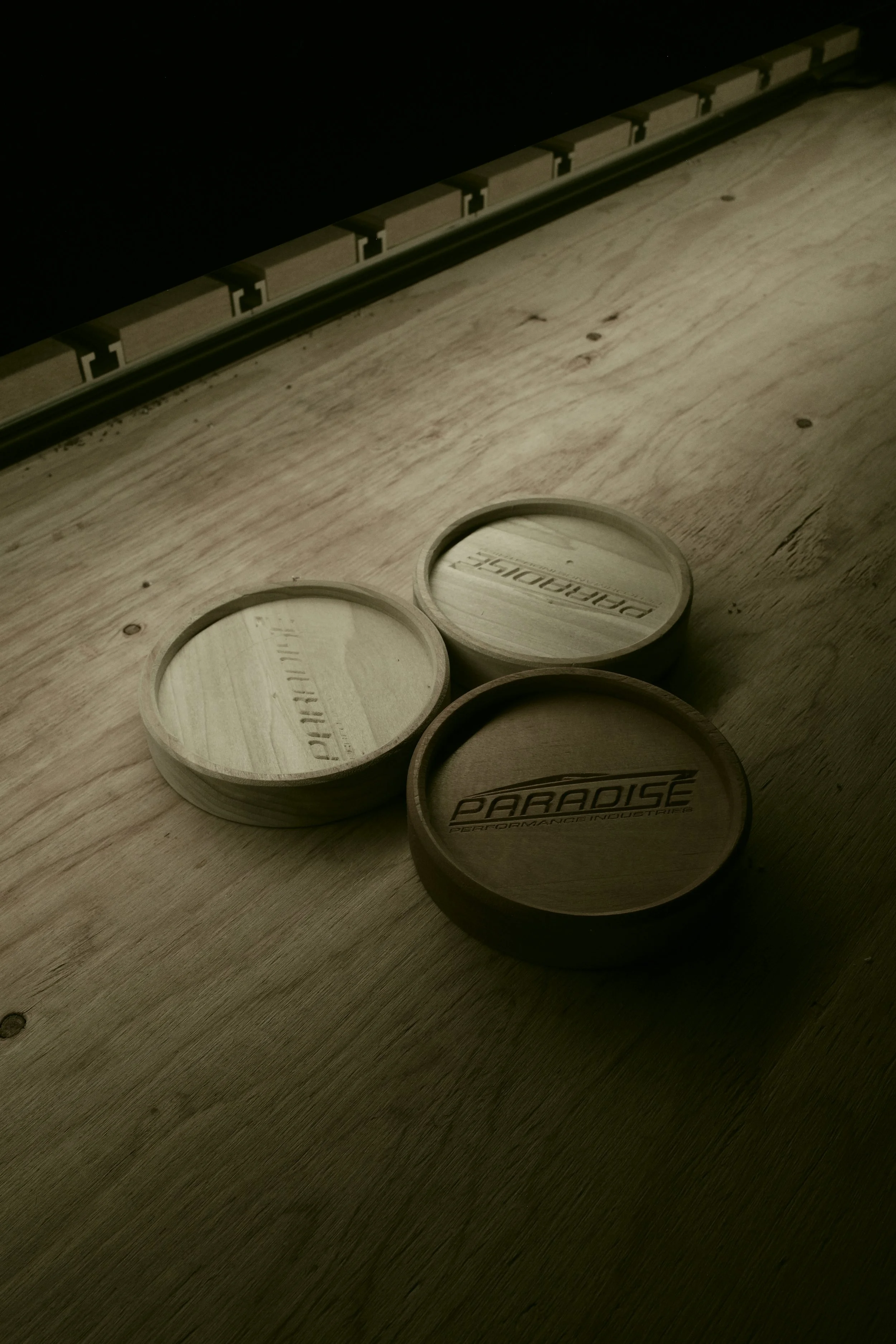 Three wooden hockey pucks on a light wooden surface near an unfinished wall, with one puck bearing the logo 'PARADISE PERFORMANCE INDUSTRIES' and another with the word 'PARTNERHIP' engraved.
