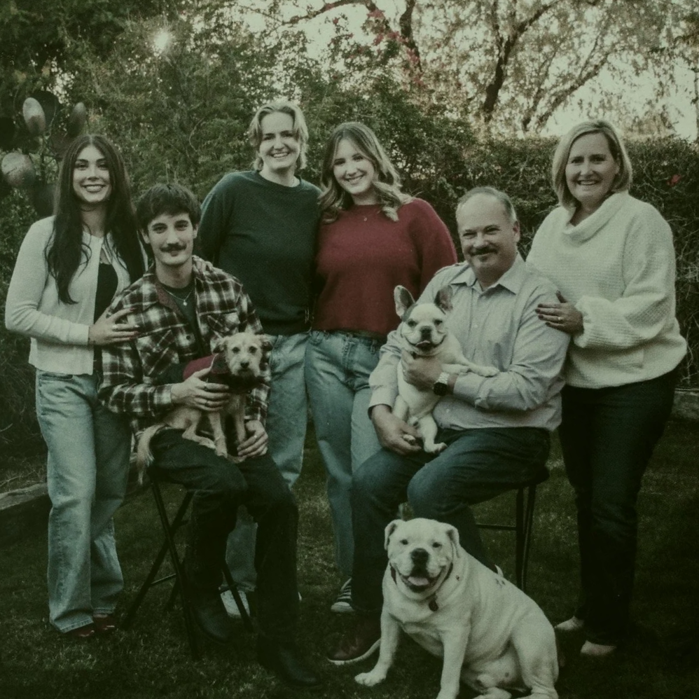 A family of seven people posing outdoors with two dogs. Four women, one man, a young boy, and a girl, all smiling. One woman and the man are sitting, holding a French Bulldog puppy, while the other three women and a young boy stand behind them. The boy is holding a small brown dog, and a large white bulldog is sitting on the ground in front of the group. The background shows trees and bushes.