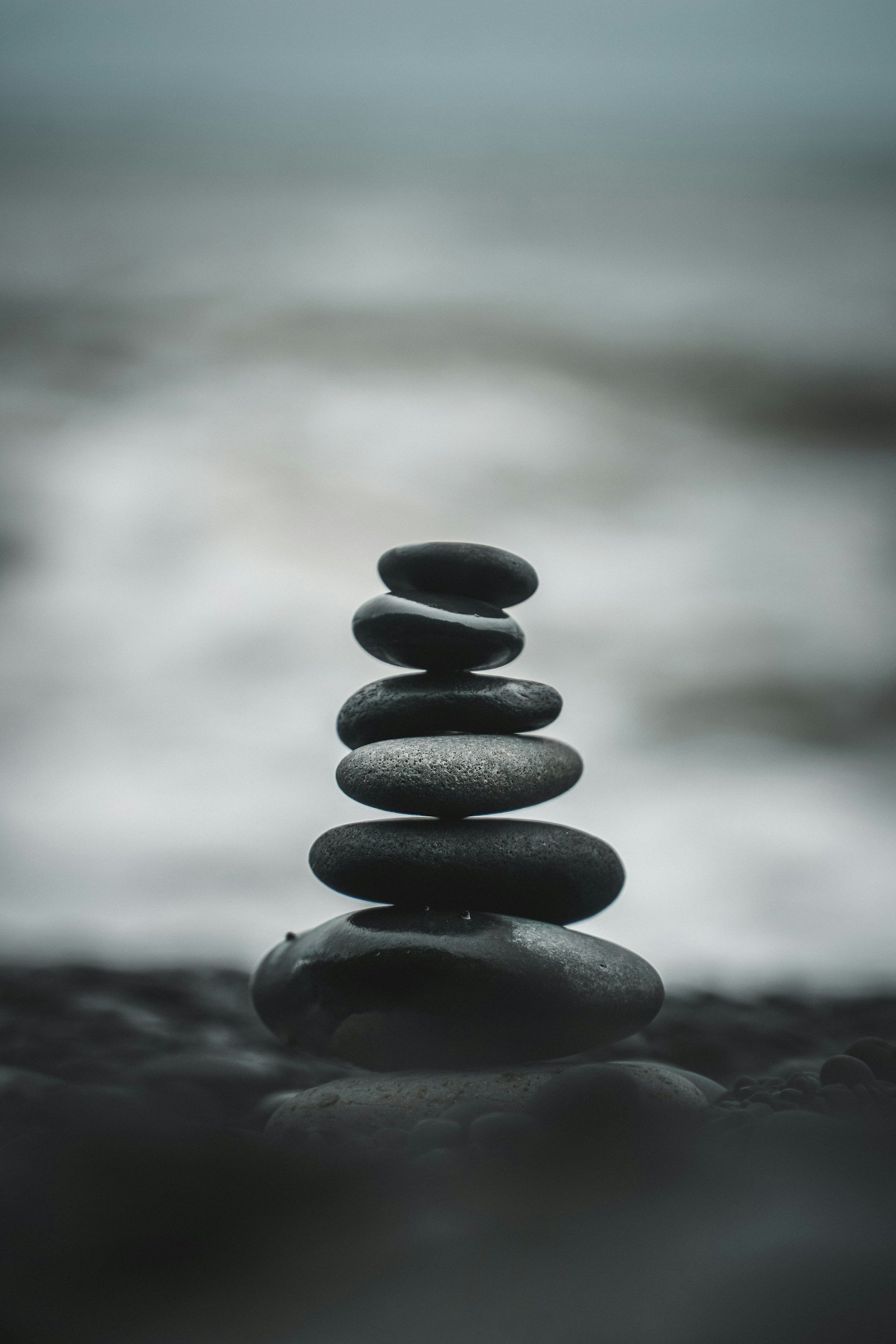 Stacked smooth black pebbles balancing on a larger stone with blurred water and sky in the background symbolizing calm.